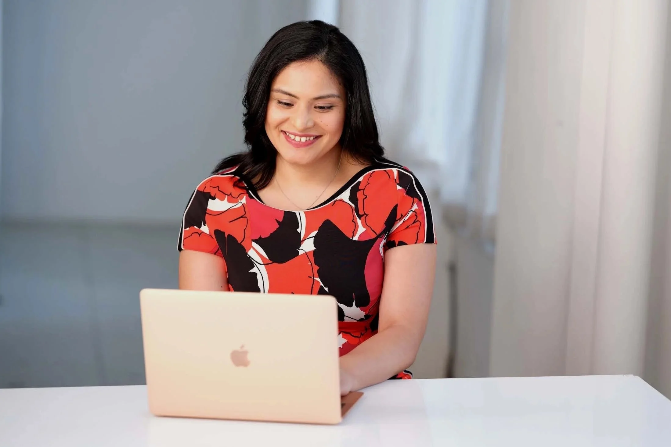 A woman with black hair smiling while working on a white MacBook at a white table in a room with soft lighting.