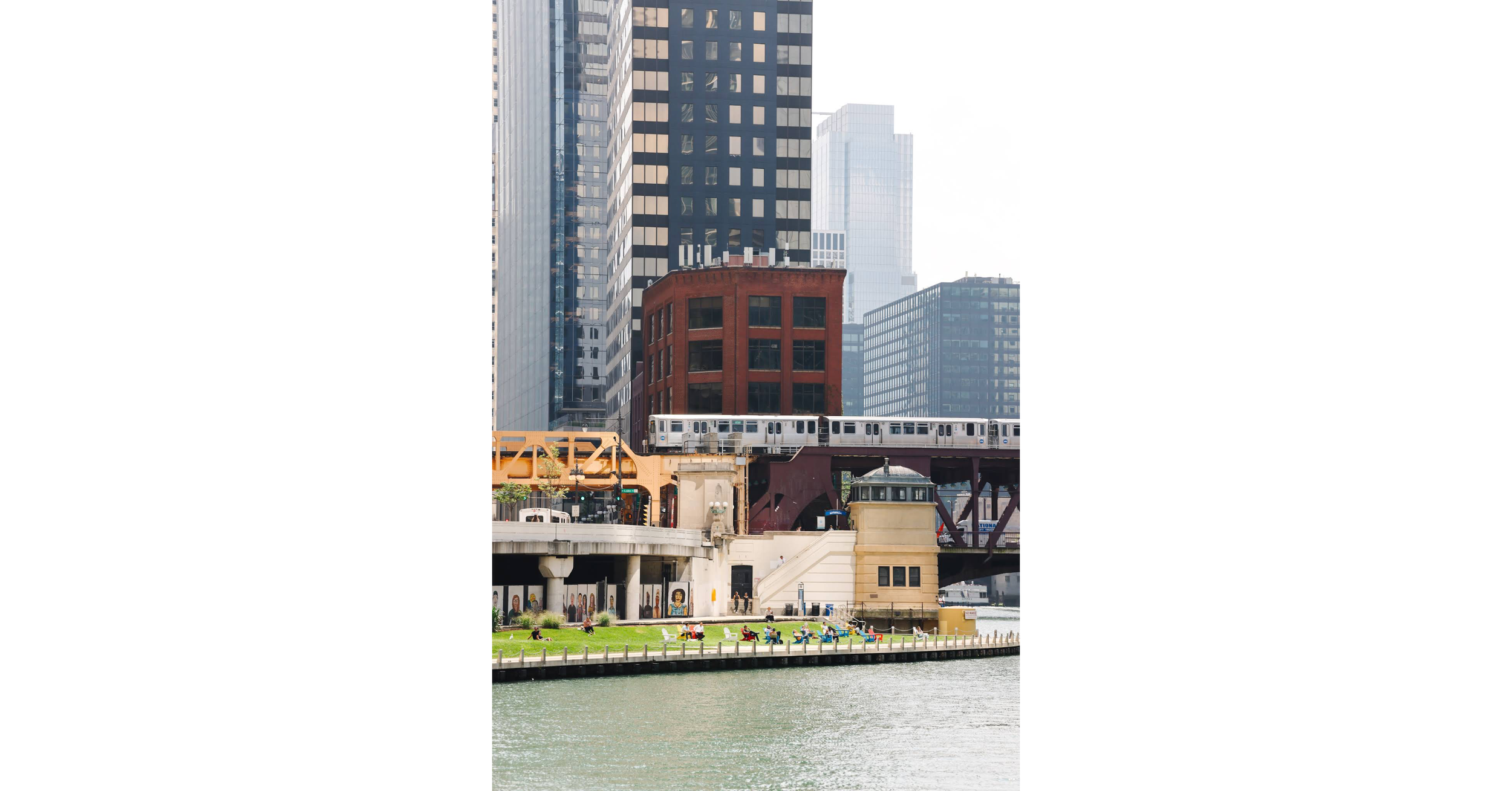 Photo of the Chicago River in the foreground with the green grass of the Riverwalk midfield, with an L train crossing a bridge and skyscrapers in the background