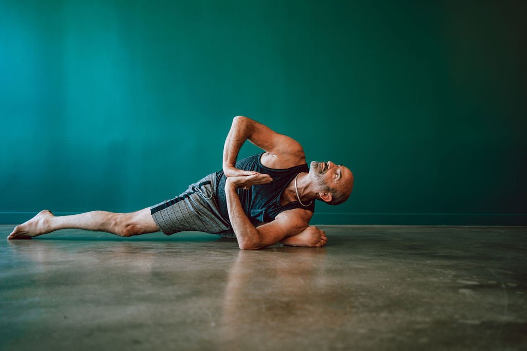 David Schlussel in a twisted pigeon pose against a green wall at Arise Yoga Oakland.