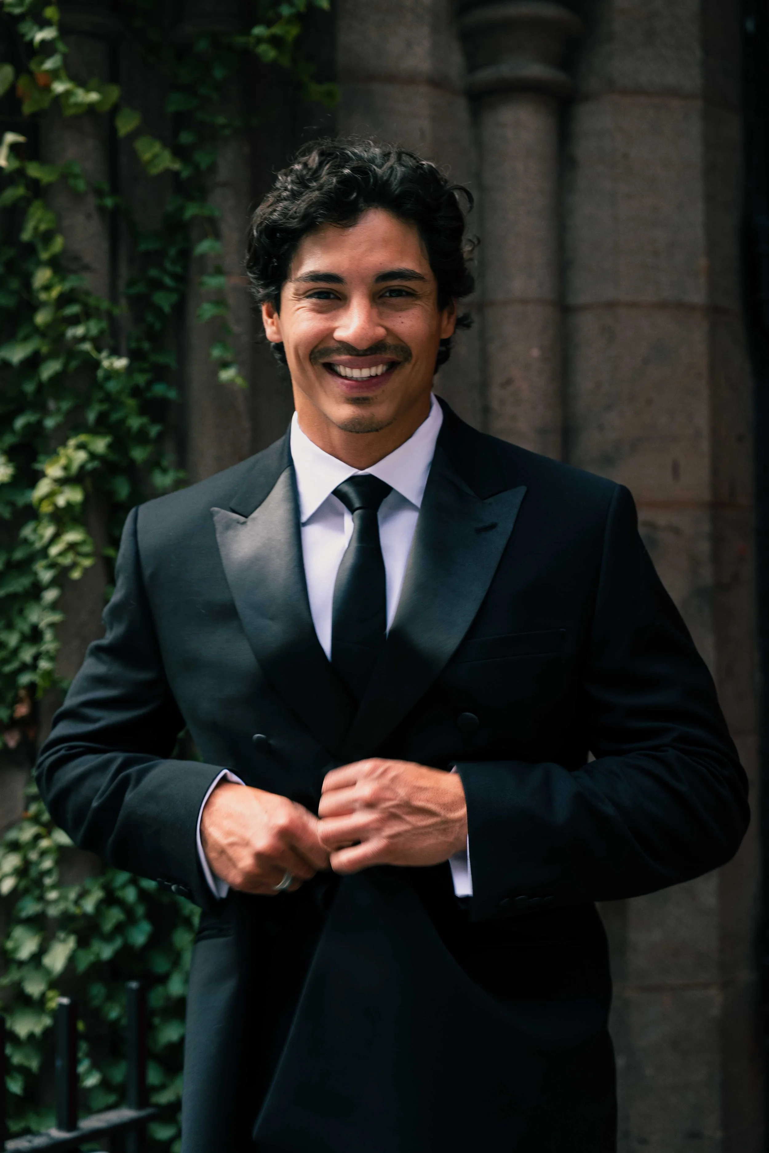 A man in a black tuxedo with a satin lapel, white shirt, and black tie, smiling while adjusting his suit, standing outside near a stone building with ivy.