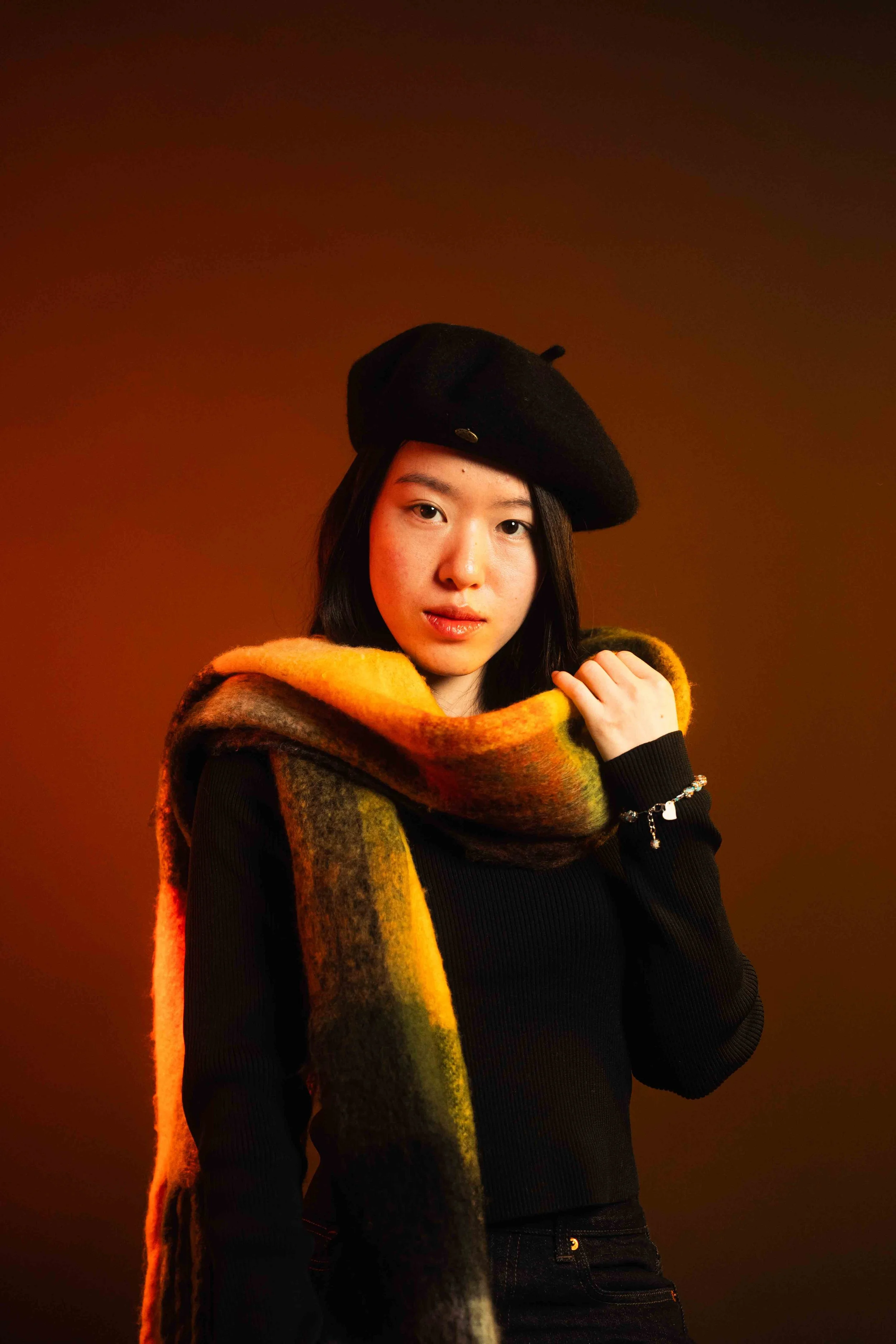 A young woman wearing a black beret and a multicolored scarf stands against a dark brown background.