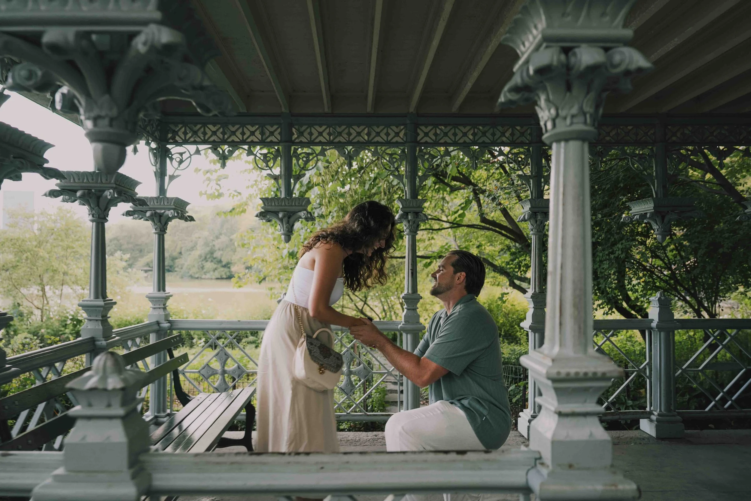A man kneeling on one knee on a gazebo, proposing to a woman in a park surrounded by greenery and trees.