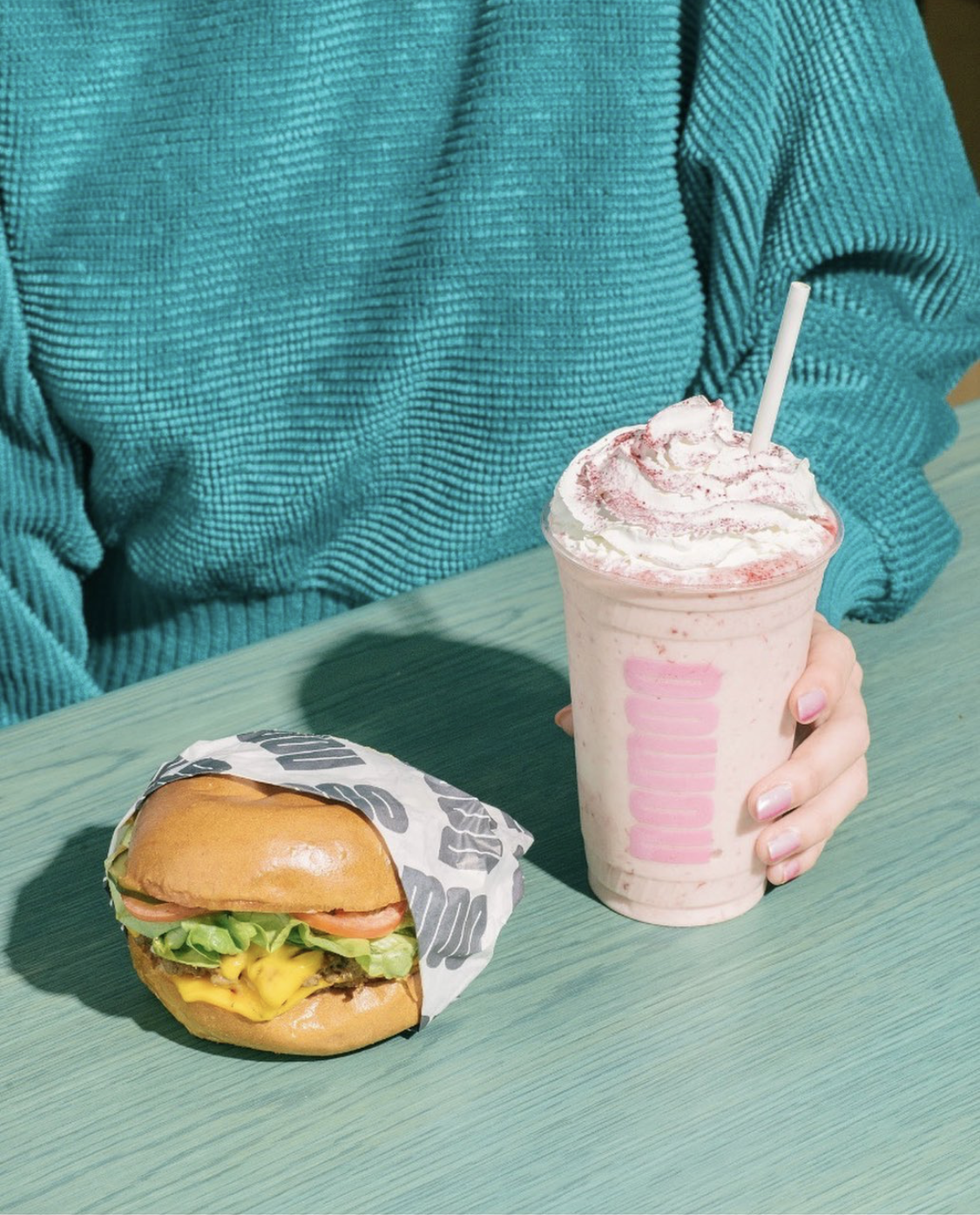A person wearing a teal sweater holding a blended strawberry milkshake with whipped cream and pink syrup on top, while a cheeseburger wrapped partially in paper sits on a green table.