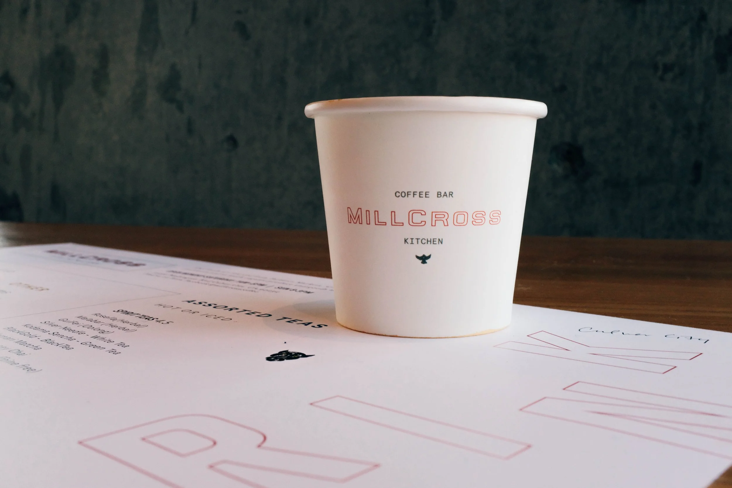 A white paper cup with the words 'Coffee Bar Millcross Kitchen' printed on it, placed on a table next to a menu or flyer with partially visible text.