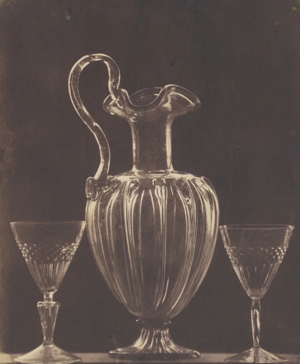 Black and white photograph of a glass pitcher with an ornate handle, flanked by two smaller glass bowls with patterned rims, against a dark background.