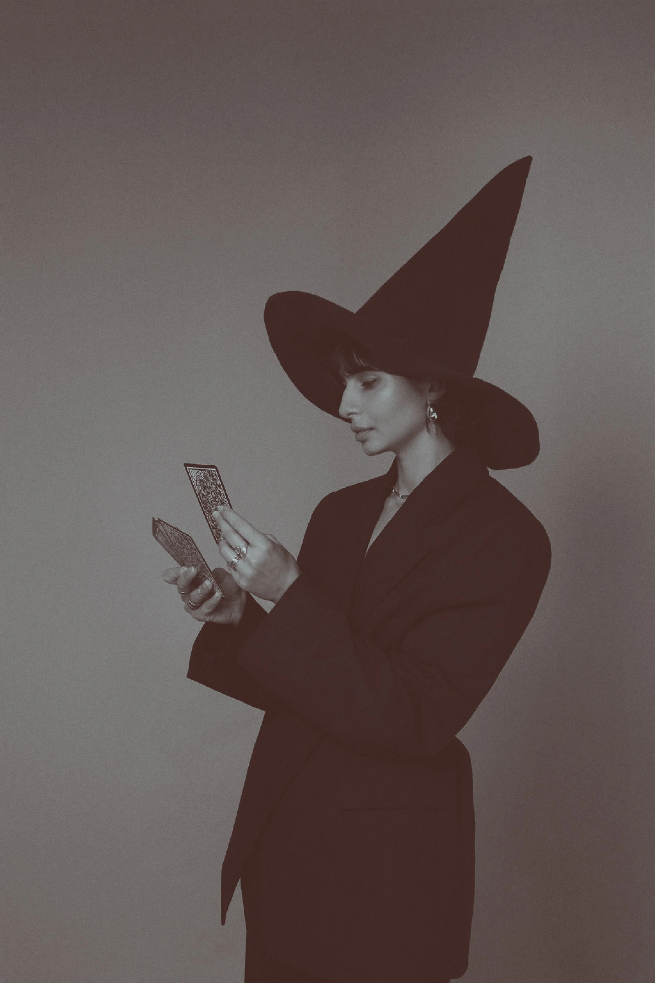 A woman dressed as a witch, wearing a large pointed hat and a suit, holding and looking at tarot cards.