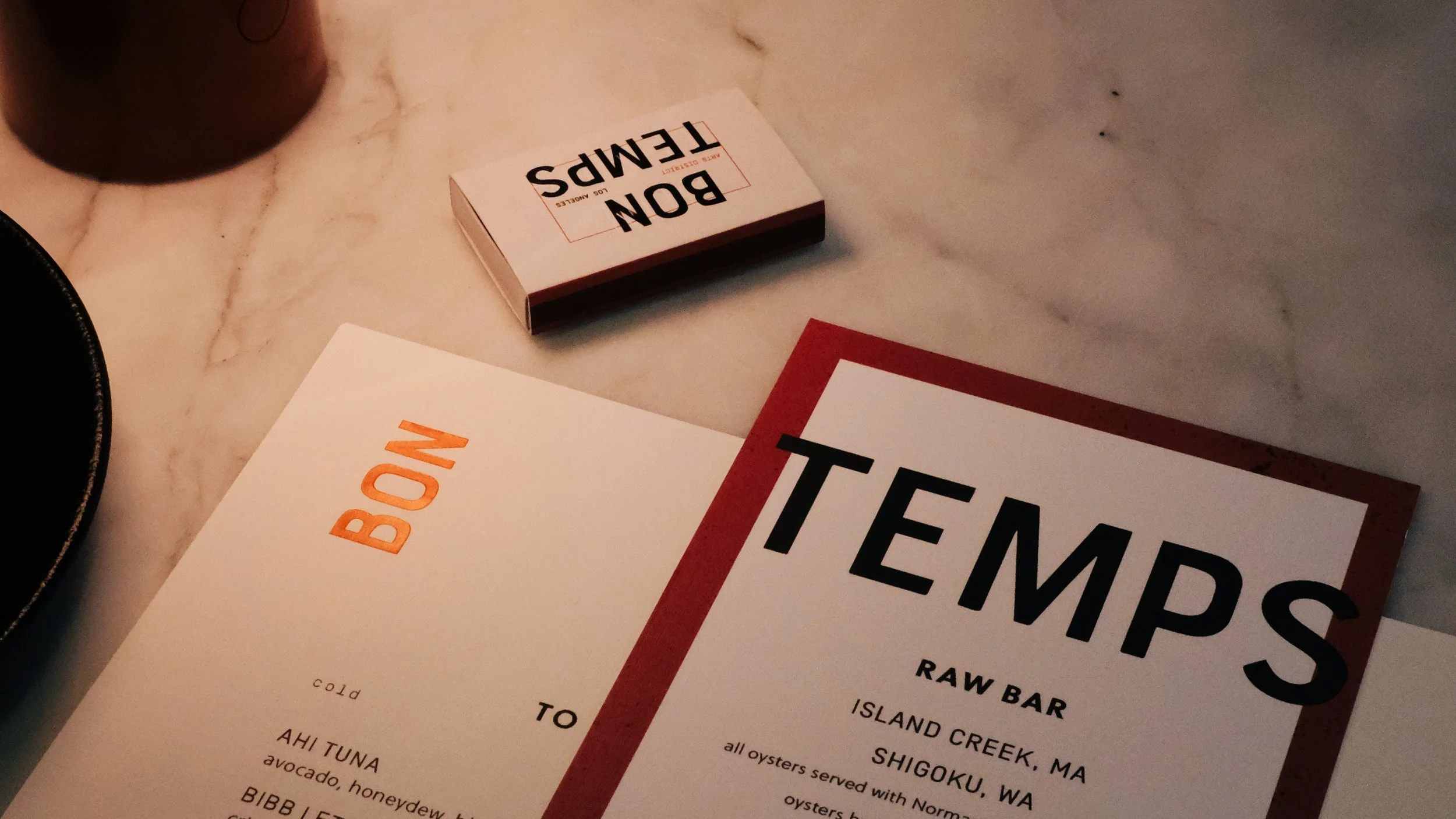A menu titled "TEMPS" and a separate menu titled "RAW" on a marble table. The TEMPS menu lists various raw bar options from Island Creek, MA, and Shigoku, WA. The BOW menu appears to be a small, folded card.