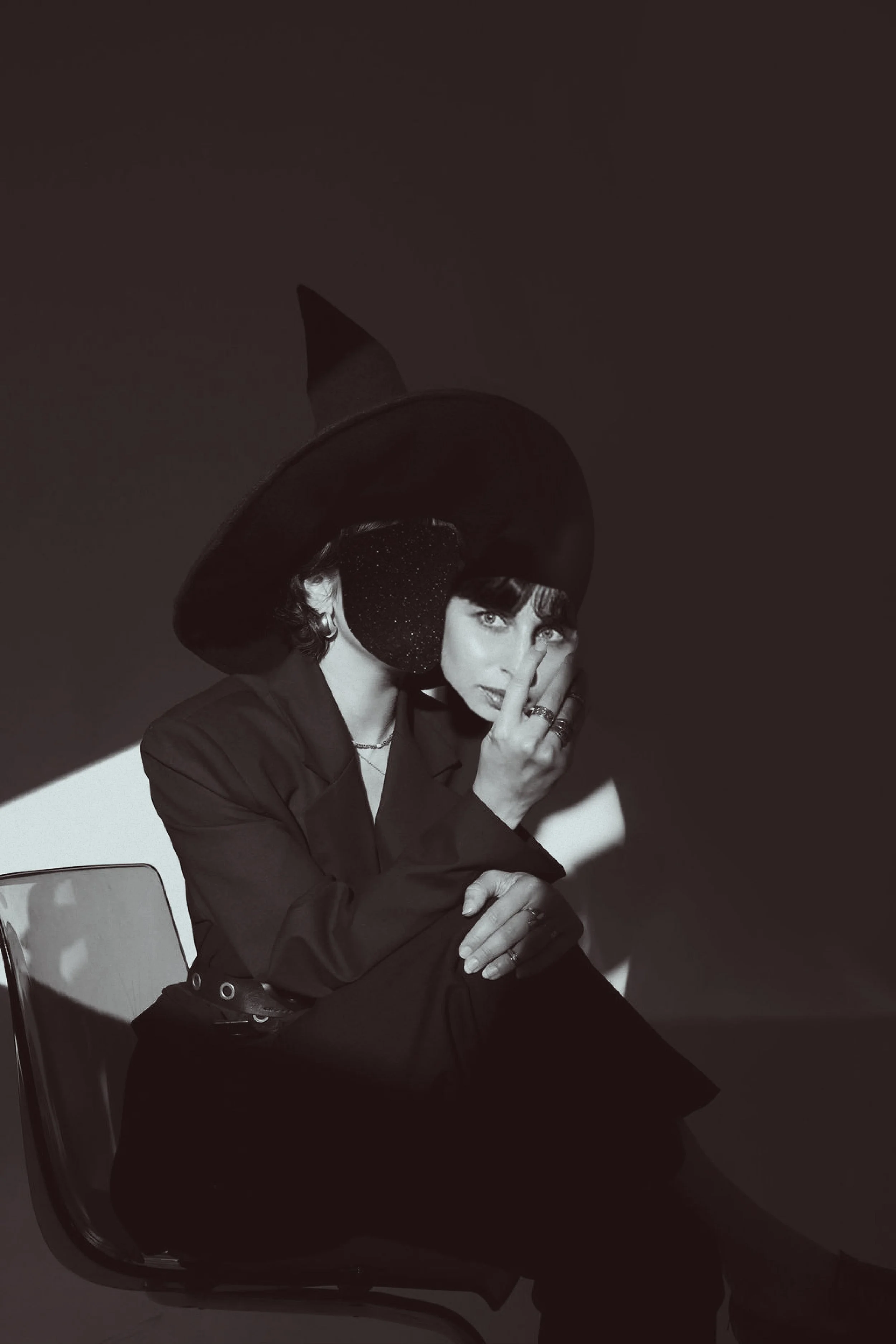 A woman in a dark blazer and makeup sitting on a chair, wearing a large witch hat with a sparkly band, covering part of her face with her hand, in black and white photo with shadowy background.