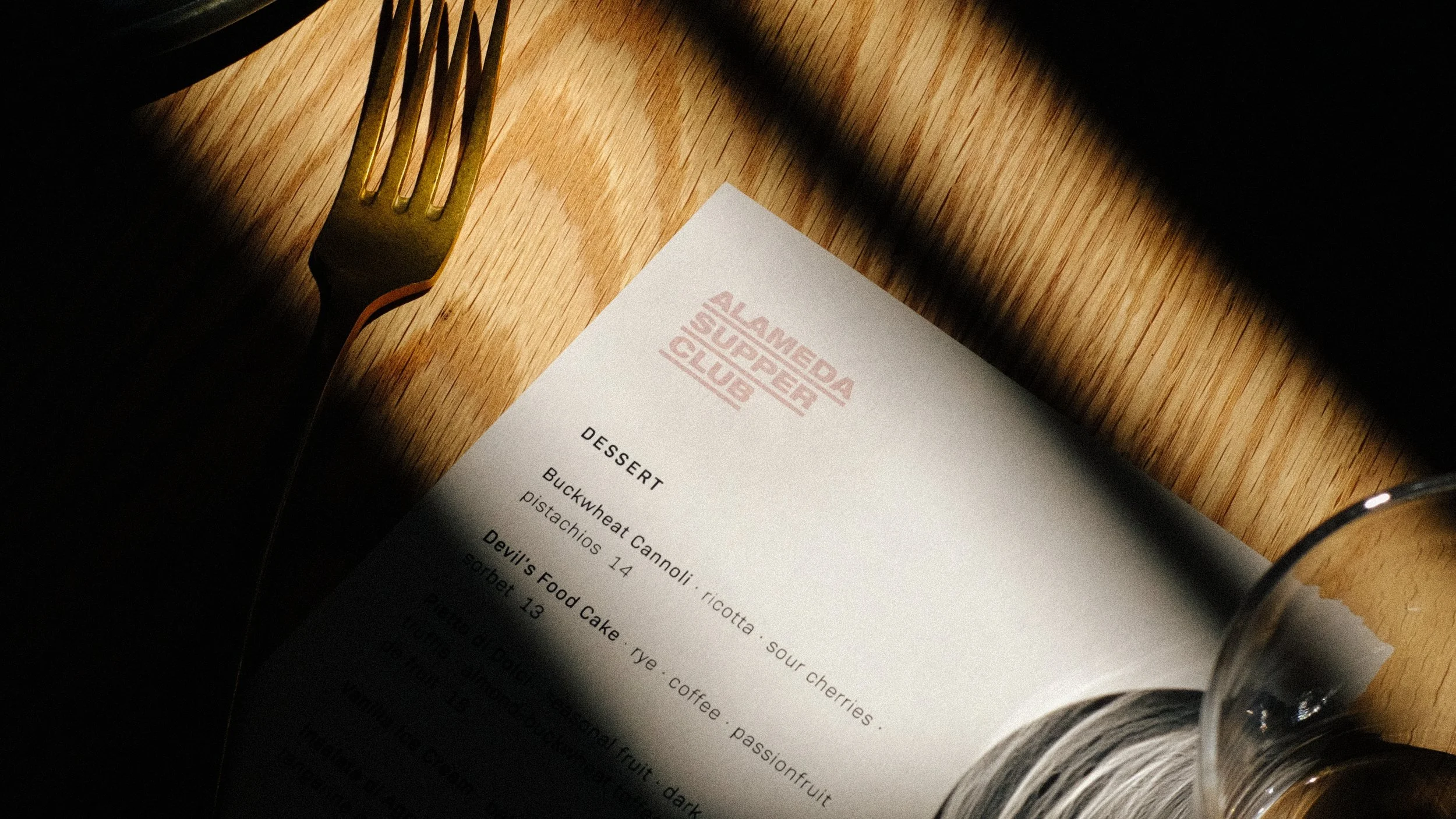 A restaurant menu on a wooden table, with a fork on the left side and part of a wine glass on the right. The menu features a logo that reads 'ALAMEDA SUPPER CLUB.'