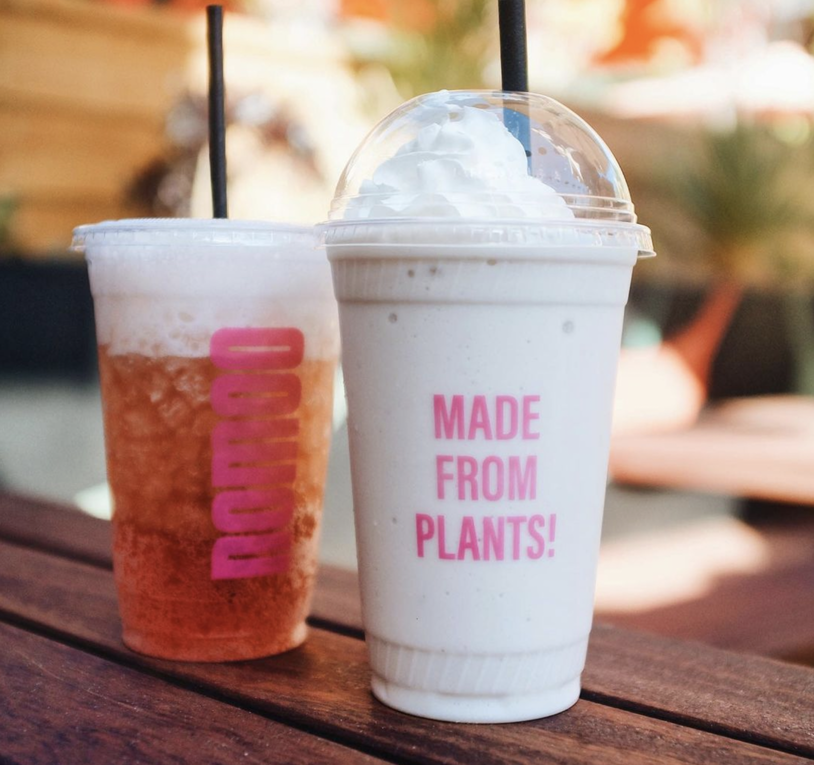 Two drinks on a wooden table, one iced tea with a black straw and whipped cream, and a creamy blended beverage with pink text saying "Made From Plants!"