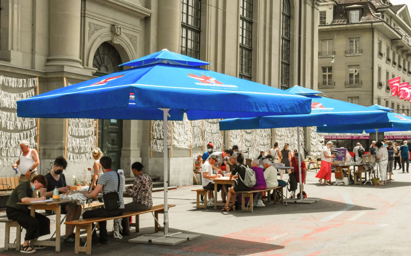 Vor der Heiliggeistkirche stehen drei Tische an denen Personen die Stoffstreifen für die Buchproduktion vorbereiten. Die Sonne scheint grell, doch die Tische sind von grossen Sonnenschirmen beschattet