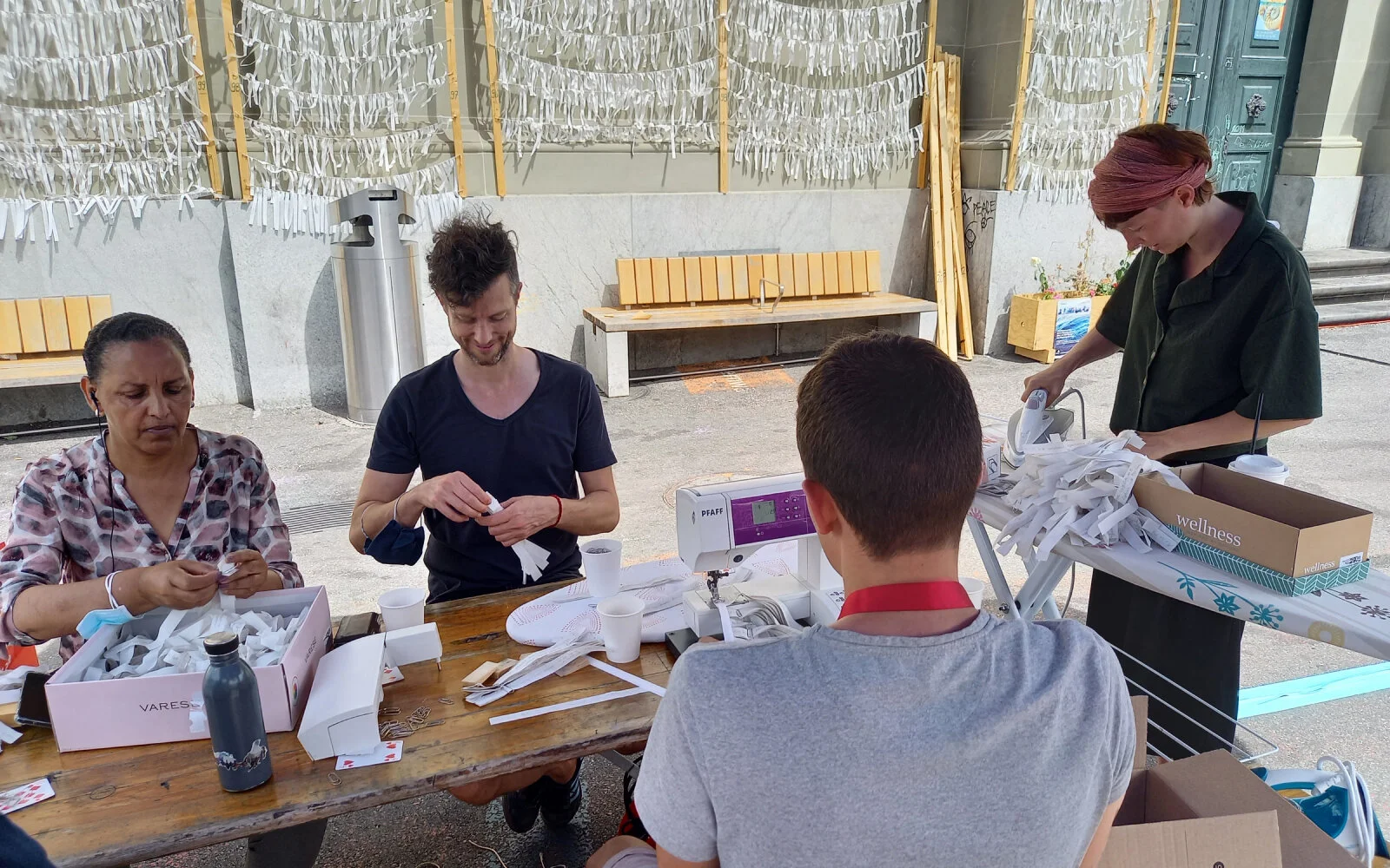 Vier Personen sitzen an einem Tisch vor der Heiliggeistkirche und bereiten die Stoffstreifen vor für die Buchproduktion. Sie bügeln und bündeln sie.