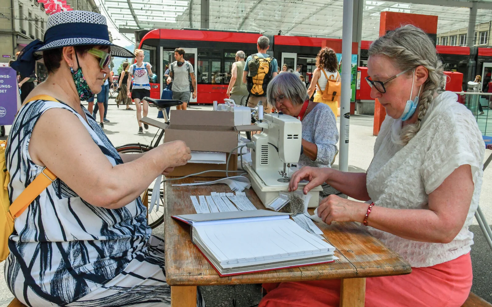 Drei Personen sitzen an einem Tisch vor der Heiliggeistkirche und bündeln Stoffstreifen. Auf dem Tisch steht eine Nähmaschine um die Streifen zusammenzunähen, sowie ein Blindband des Buches als Vorlage
