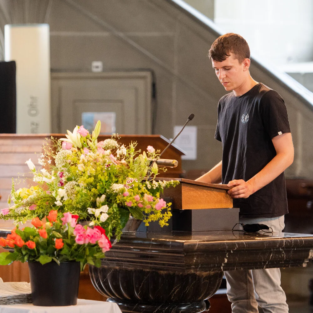 Ein junger Mann steht am Podium in der Heiliggeistkirche und liesst von der «List of Deaths». Vor ihm steht ein Blumenarrangement.