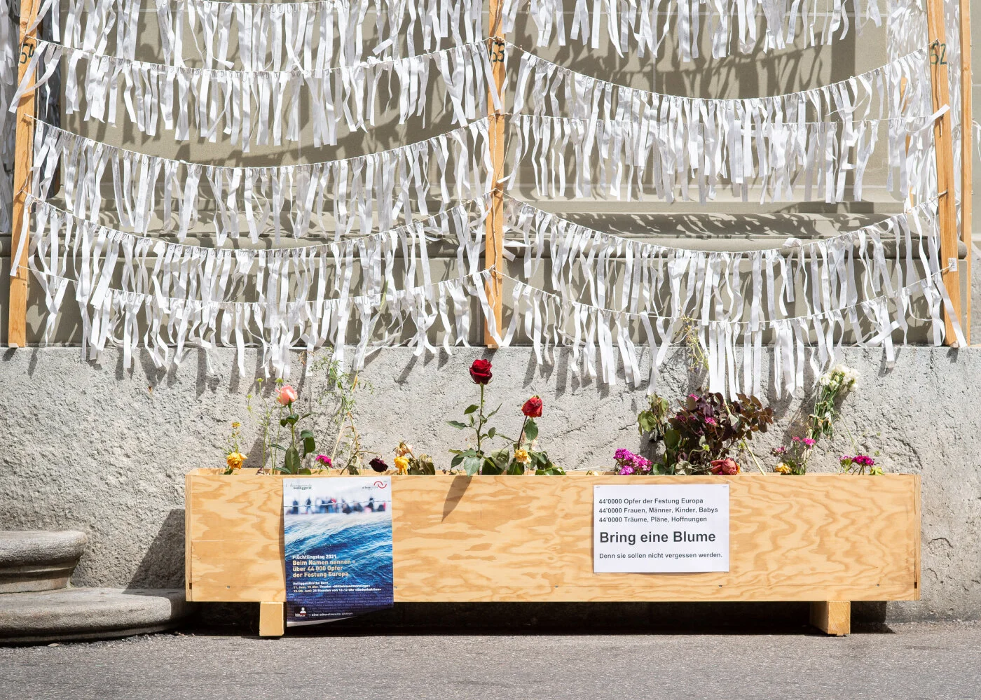 An der Fassade der Heiliggeistkirche hängen Stoffstreifen dicht neben- und übereinander. Darunter ein gefäss aus Holz, in dem verschiedene Blumen stehen und eine Aufschrift «Bring eine Blume»