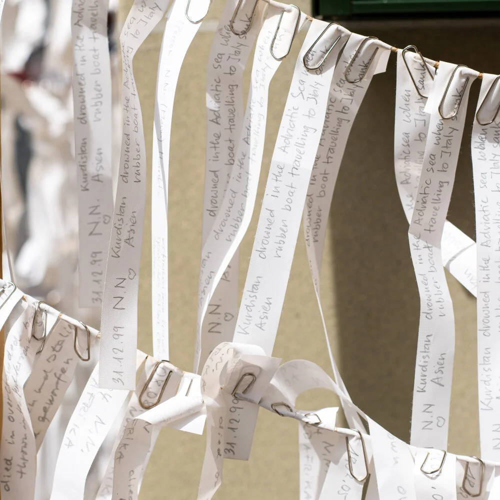 Nahaufnahme einzener hängender Stoffstreifen. Die Angaben der Verstorbenen sind in feiner Handschrift darauf vermerkt