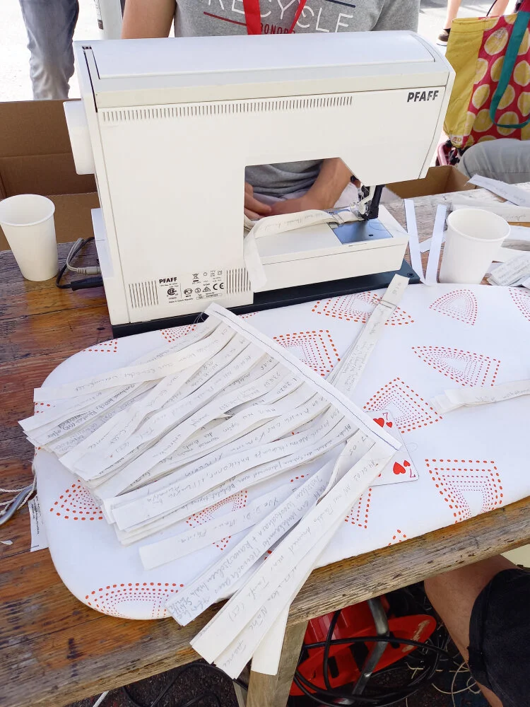 14 Bündel à 10 glattgebügelten Stoffstreifen wurden mit der Nähmaschine zu einer Einheit zusammengenäht. Dieses Stück liegt gleich neben einer Nähmaschine auf einem Tisch.