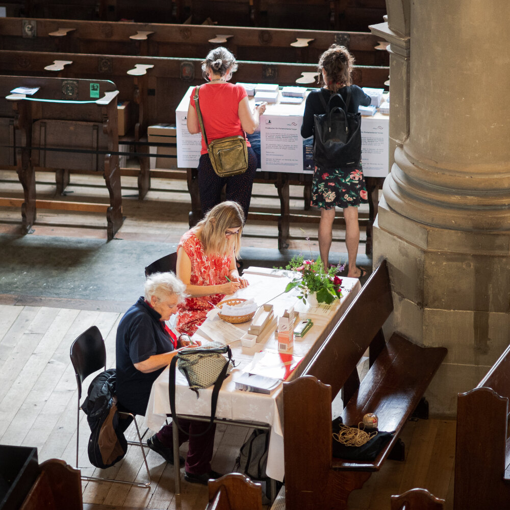 Zwei Menschen sitzen in der Heiliggeistkirche an einem Tisch und beschreiben Stoffstreifen, dahinter stehen zwei weitere Personen, die Postkarten für die Paramentsabgeordneten schreiben.