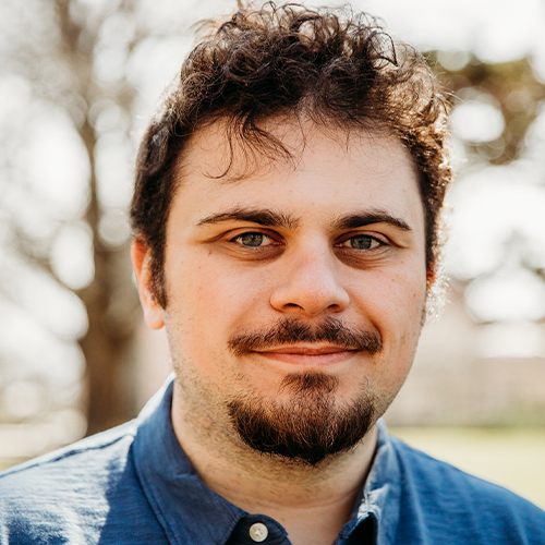 Close-up of a young man with curly brown hair, a mustache, and a goatee, wearing a blue button-up shirt, outdoors with blurred trees in the background.