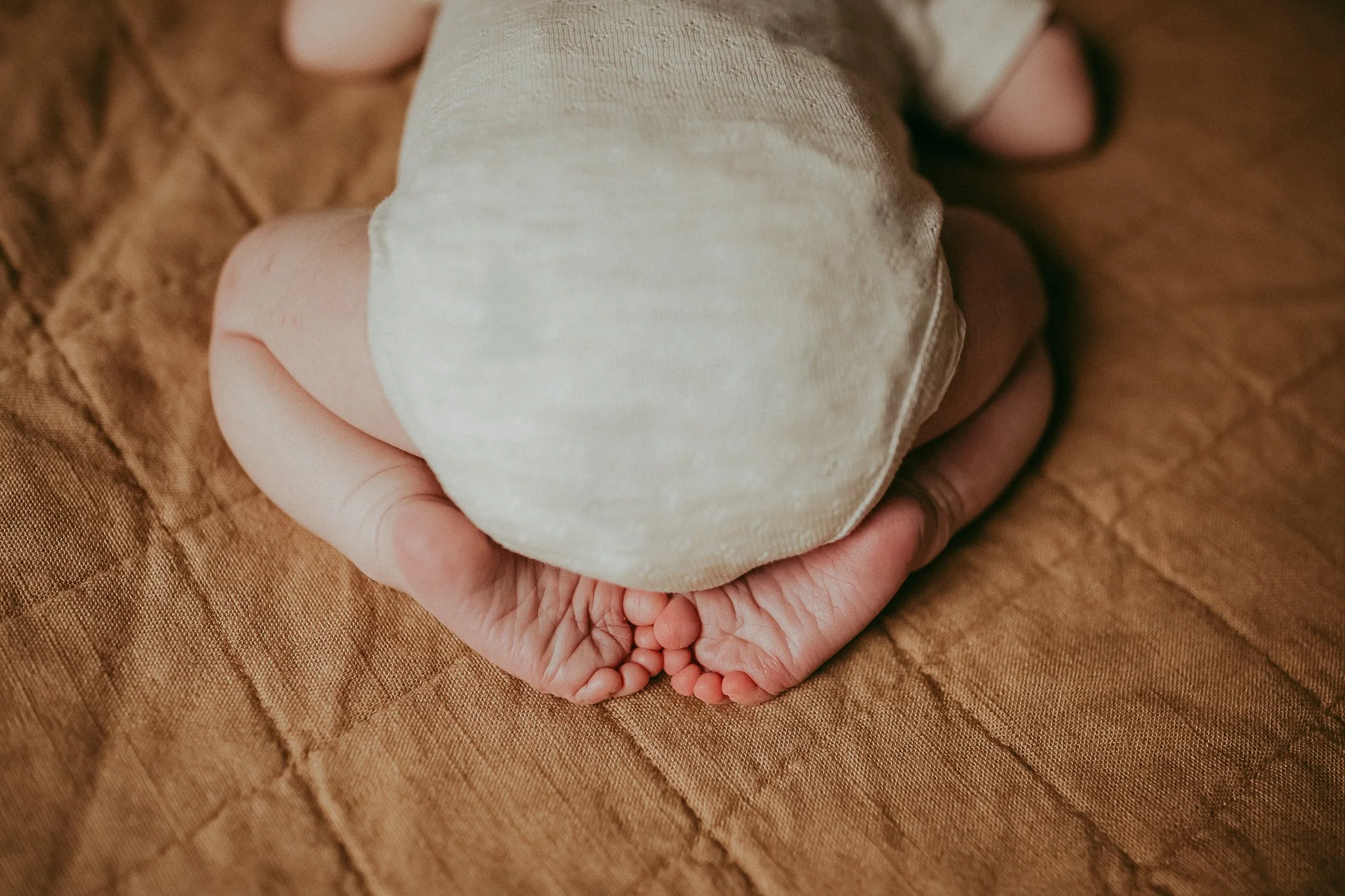 Close-up of a child's hand holding their feet, with the child wearing beige shorts, on a brown quilted fabric surface.