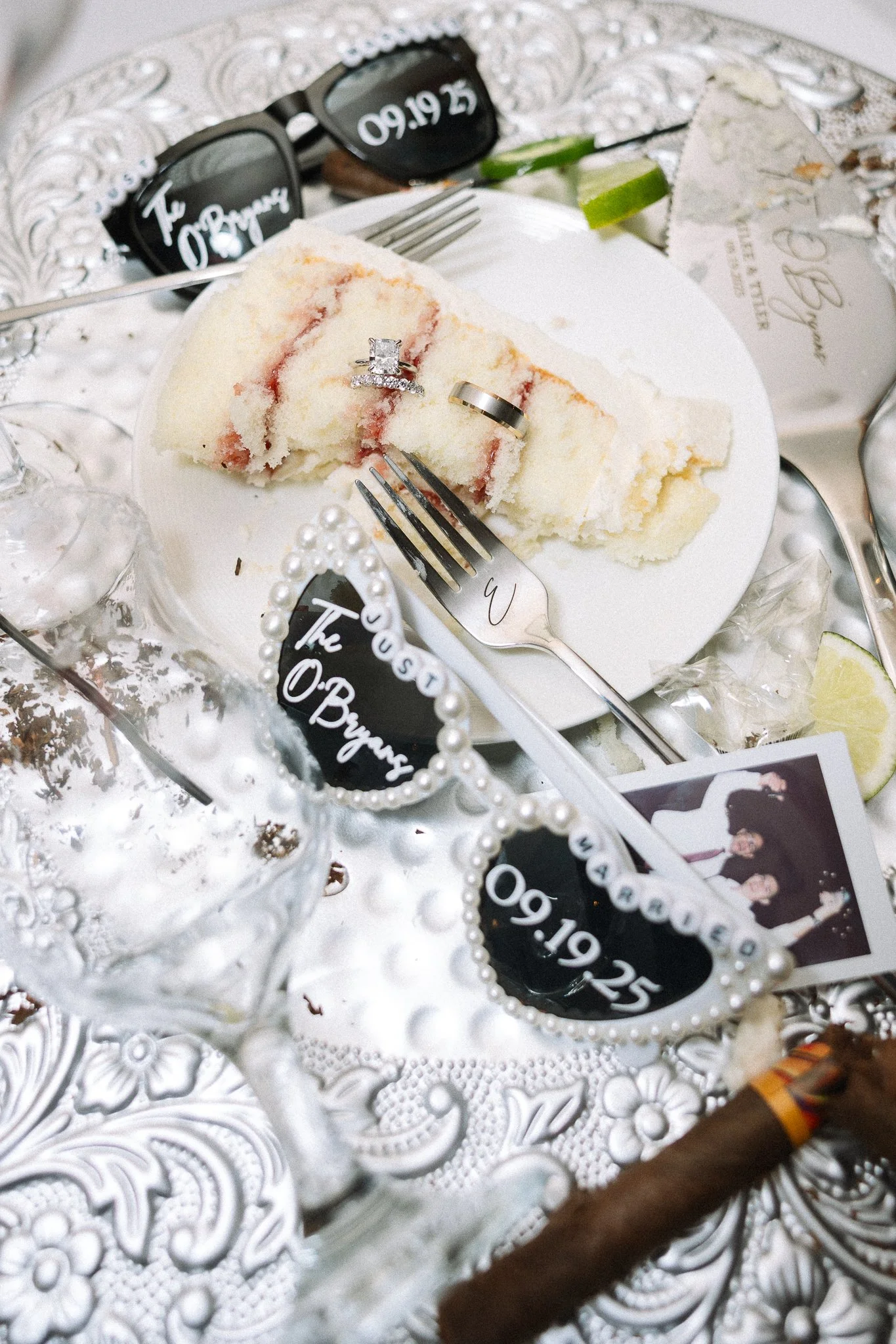 Wedding cake slice with two rings on top, surrounded by sunglasses, photos, lime wedges, pearls, and a glass of water on a decorative silver table.