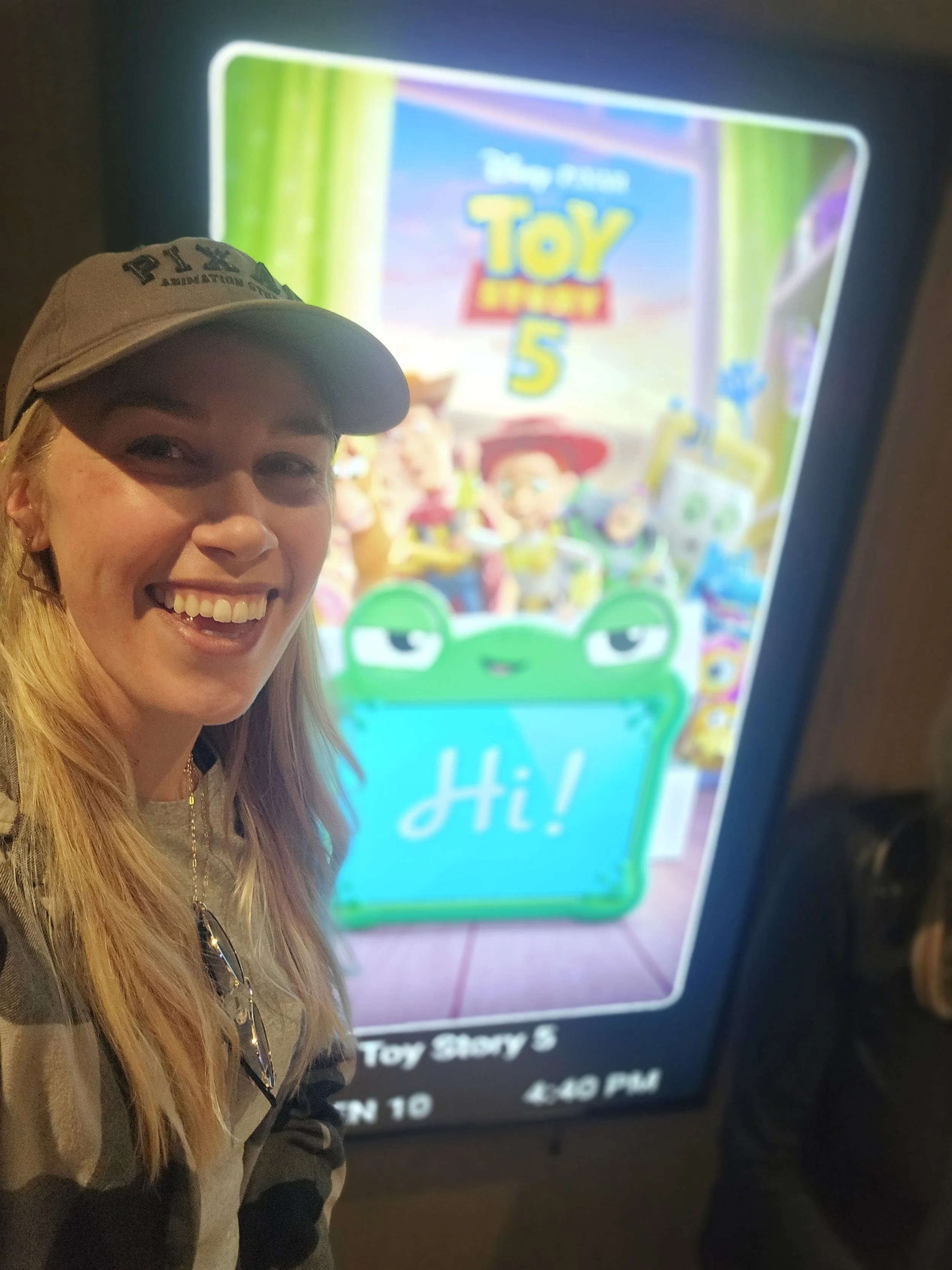 A smiling woman with long blonde hair and a beige cap taking a selfie in front of a screen displaying Toy Story 5 poster.