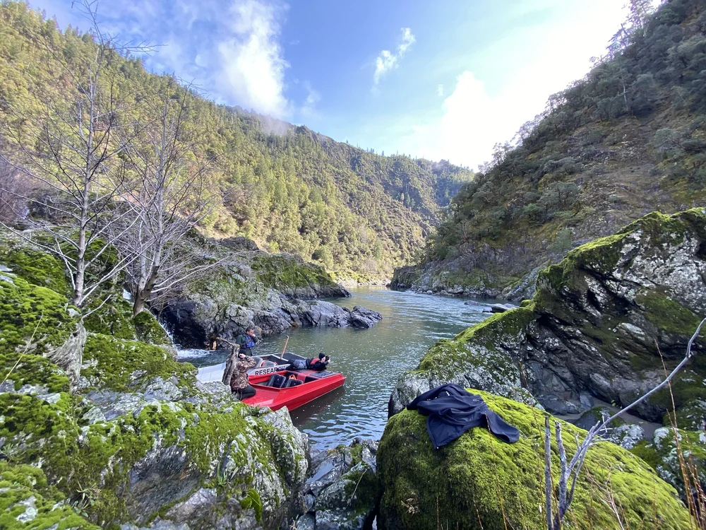 A completed red jet boat beside a moss-covered rock wall on the river