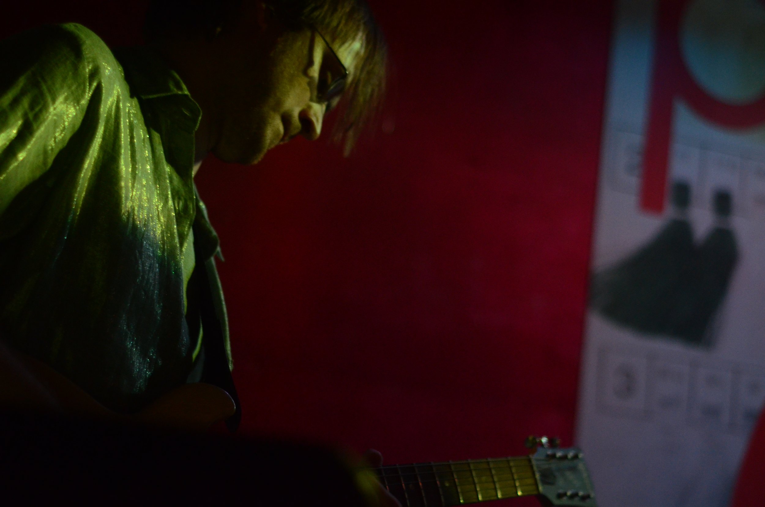 A guitarist wearing a silver shirt plays his instrument in front of a red background