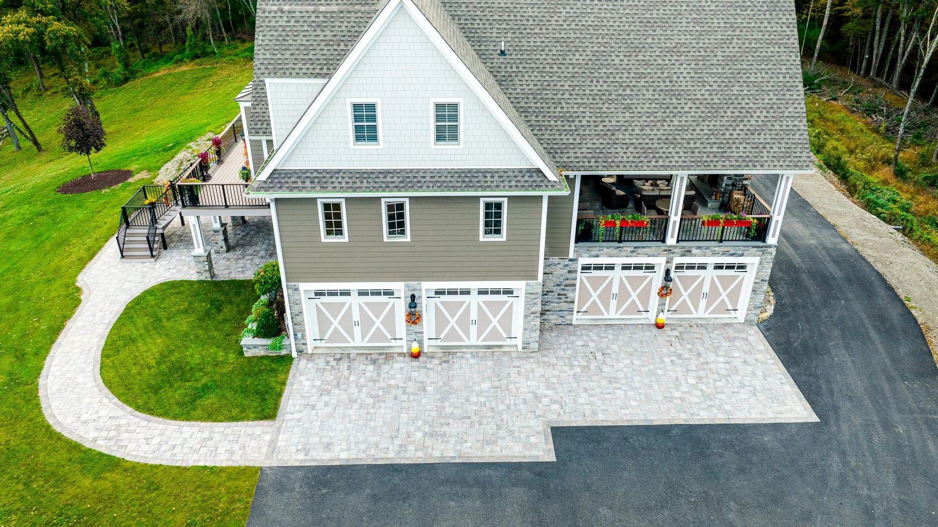 A top-down aerial view of a two-story house with a gray shingled roof, beige and green siding, a large covered porch, and four garage doors, situated on a property with a curved driveway, a grassy lawn, and a wooded area in the background.