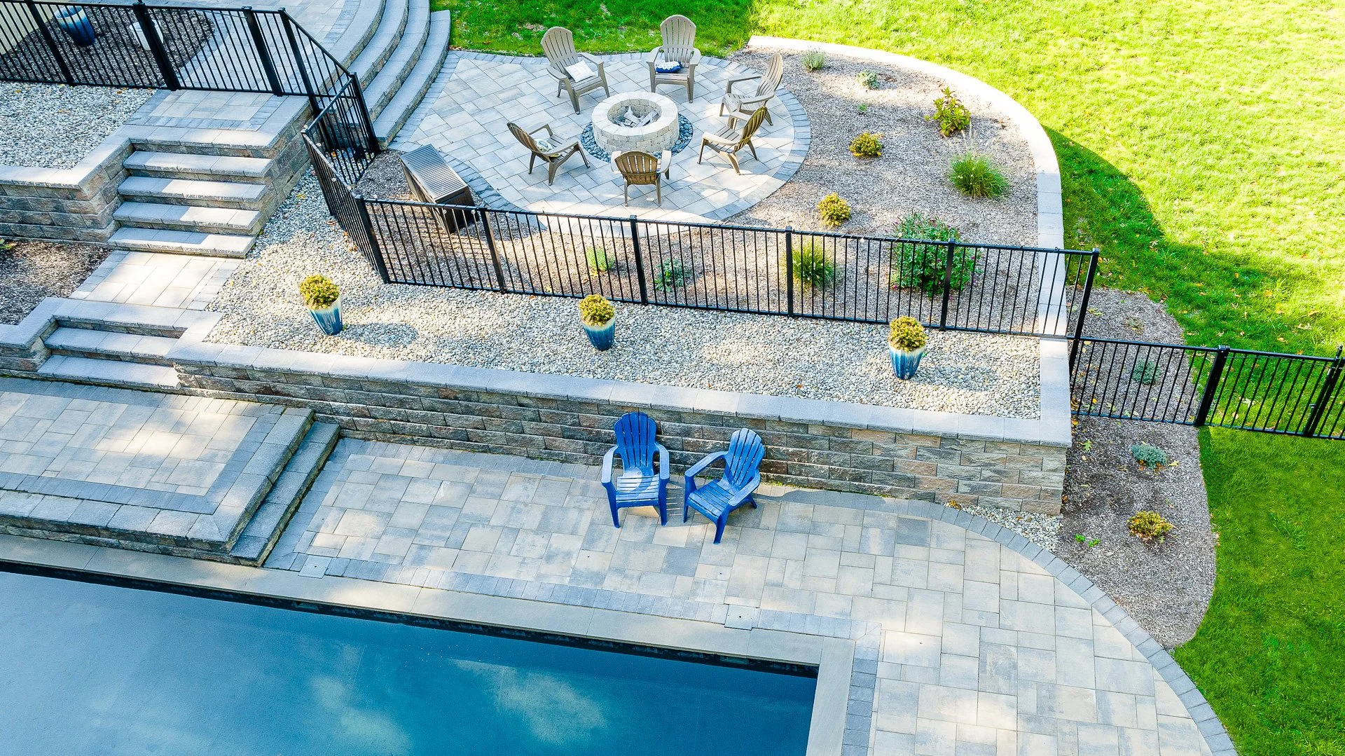 A backyard patio area viewed from above, with stone steps leading down to a paved space. The patio has a fire pit surrounded by six chairs, and there are two blue Adirondack chairs nearby. The area is enclosed by a black metal fence, with landscaped 