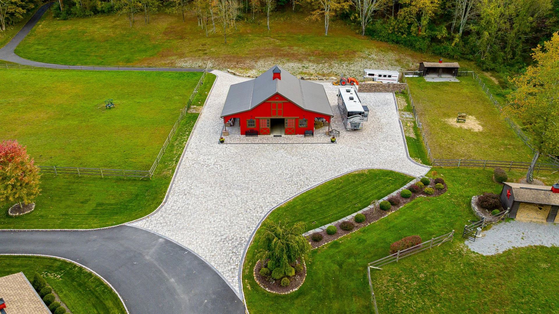 Aerial view of a red barn with gray roof, white trim, and a large paved driveway surrounded by green lawns, trees, and fencing. There are benches, a tractor, a trailer, and a small shed nearby.