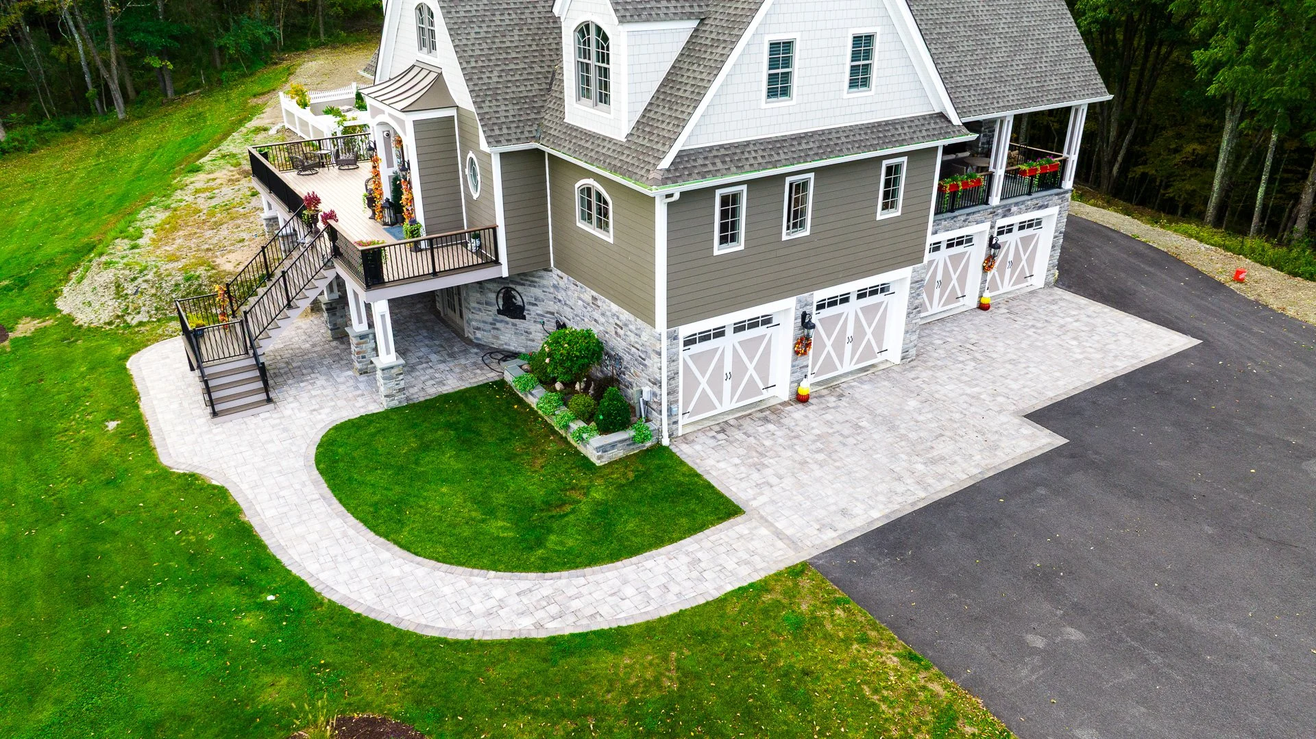 An aerial view of a modern house with a gray and white exterior, multiple garages, and a landscaped front yard with a curved pathway. Showcasing the hardscaping work done with pavers.