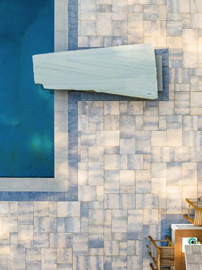A swimming pool with blue water, surrounded by beige and gray pavers. A large pale stone diving board is near the pool, with two wooden chairs and a small table with a potted plant in the corner.