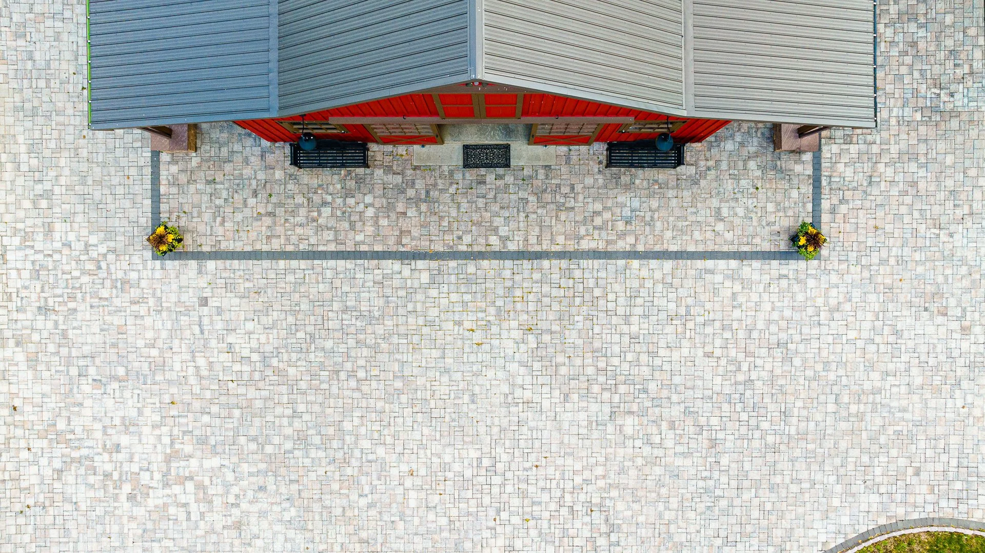 Top-down view of a barn with a gray and red roof, flanked by two benches, surrounded by a stone paver courtyard with two flower pots on opposite corners.