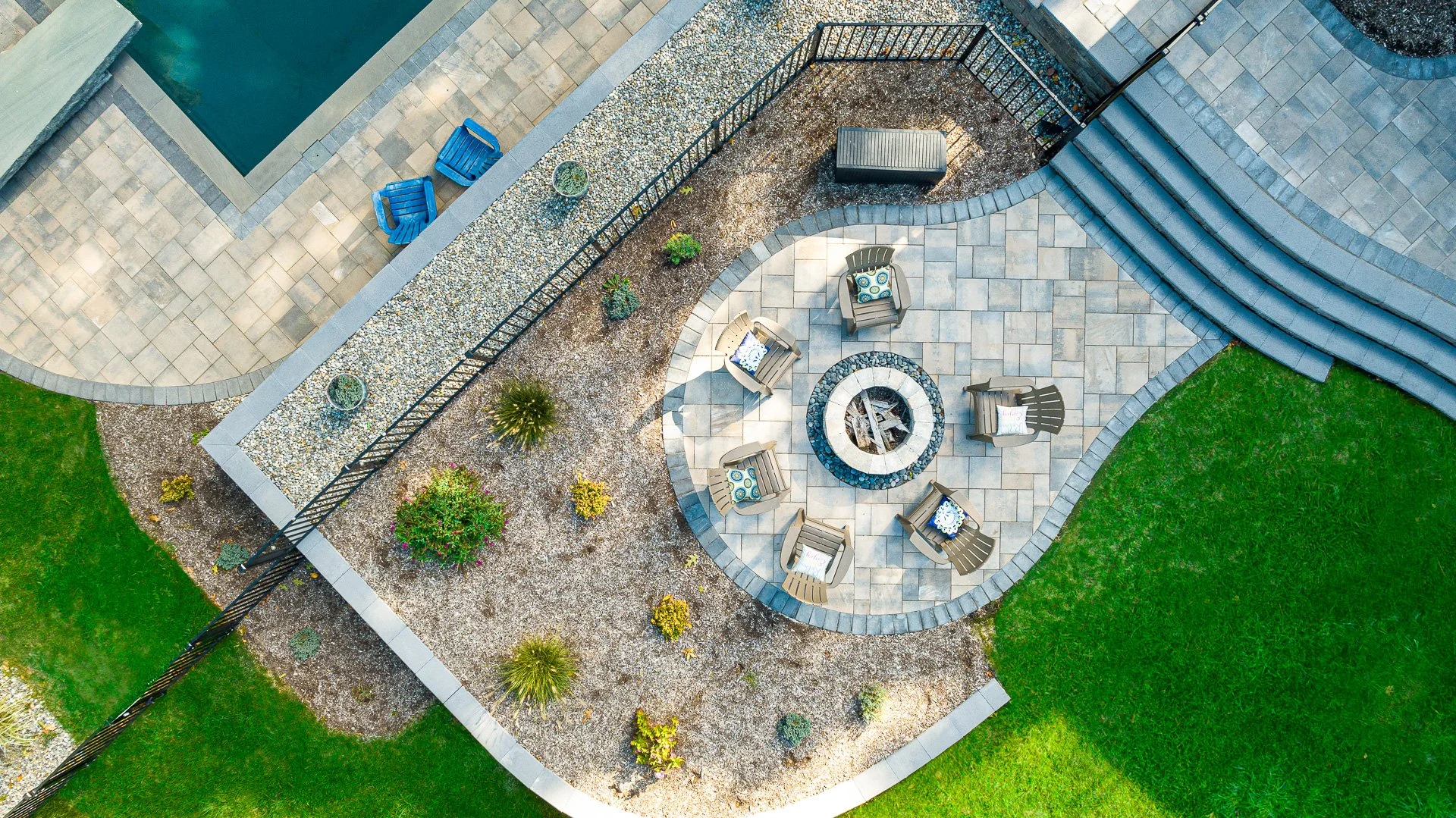Aerial view of a backyard patio with a fire pit, surrounded by six Adirondack chairs, and decorated with plants, pillows, and outdoor furniture, bordered by grass and a fence.
