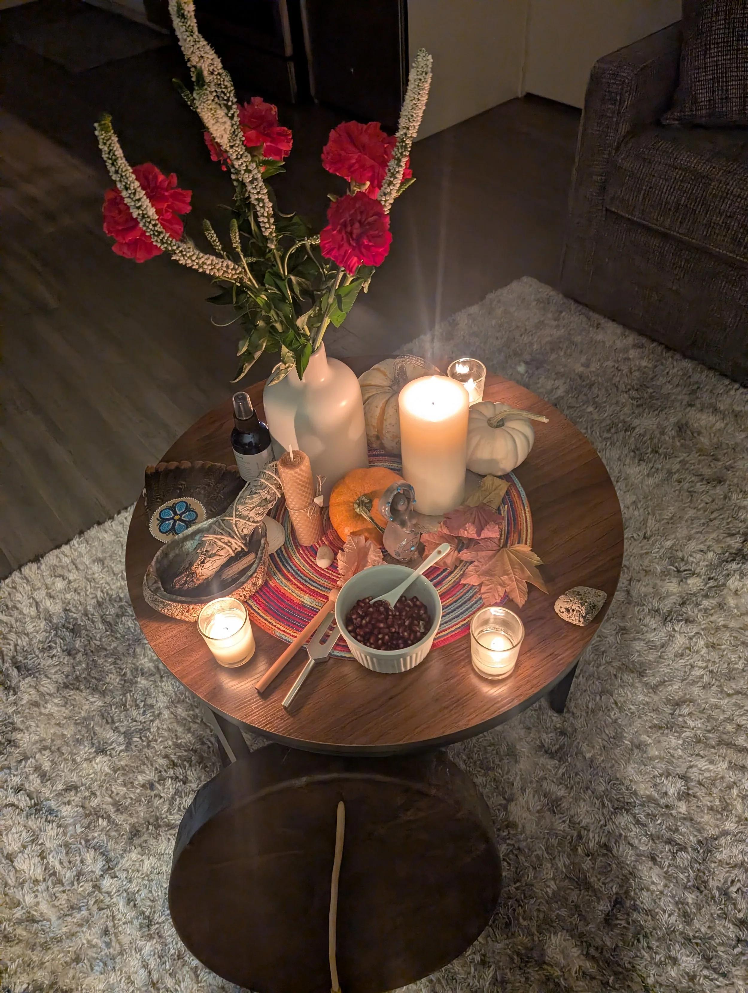 Sacred Shamanic Altar -Fall Season theme with a bouquet of red and white flowers, white and orange pumpkins, candles, autumn leaves, a bowl of cranberries, a small basket, all on a colorful round placemat, with candles representing the 4 directions.