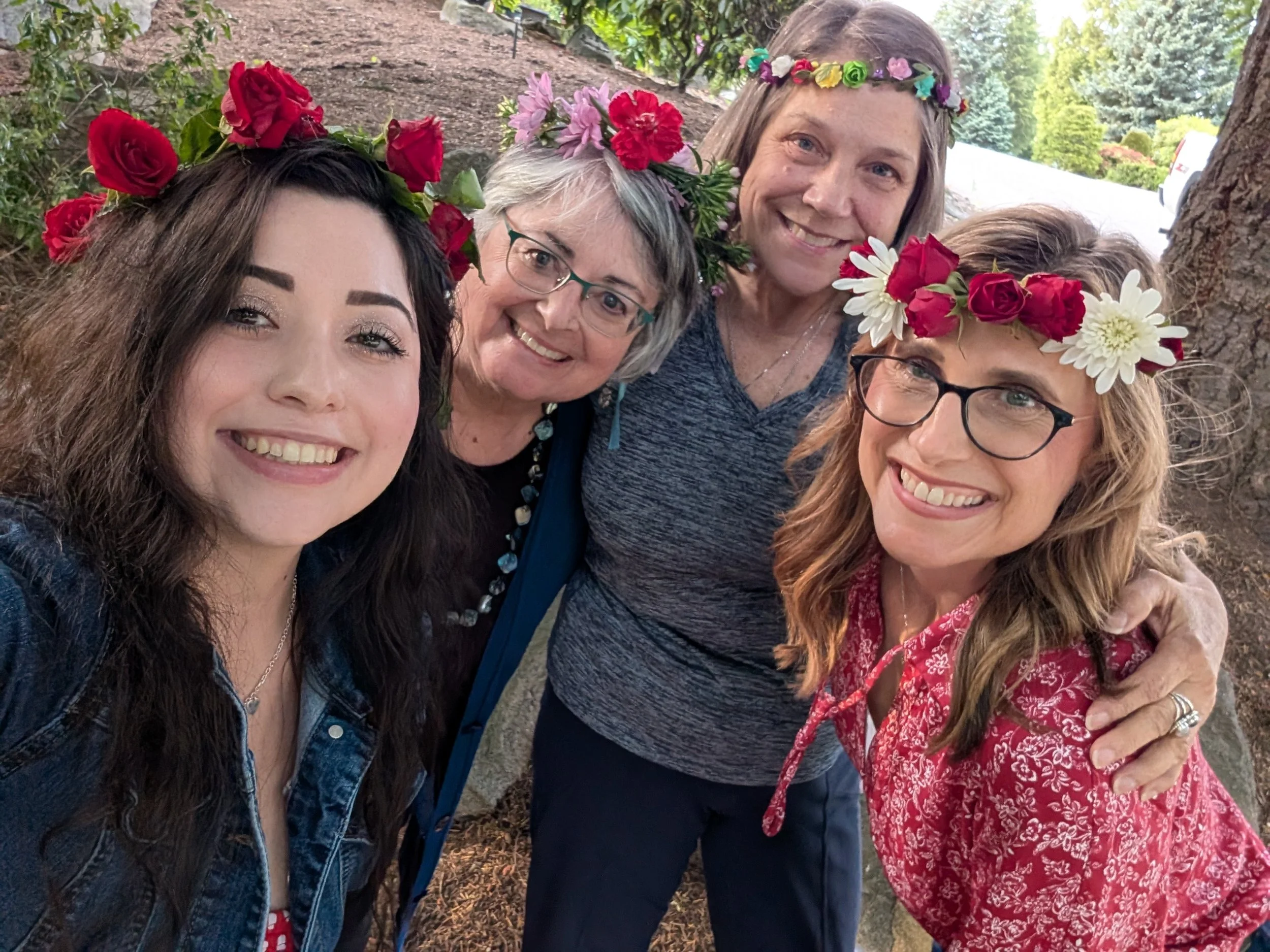 Four women with flower crowns smiling outdoors, standing close together in front of a tree and greenery.