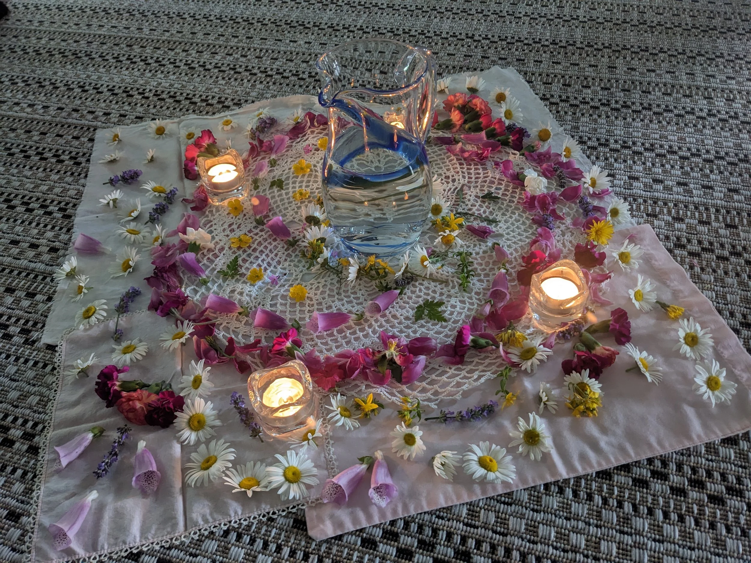 A Flower Mandala decorative arrangement on a lace cloth with a glass pitcher of water, surrounded by candles, flowers, and petals, on a textured carpet.