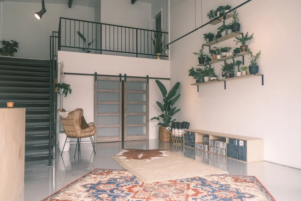 The empty studio interior: a double-height white room with a staircase, hanging plants and a patterned rug