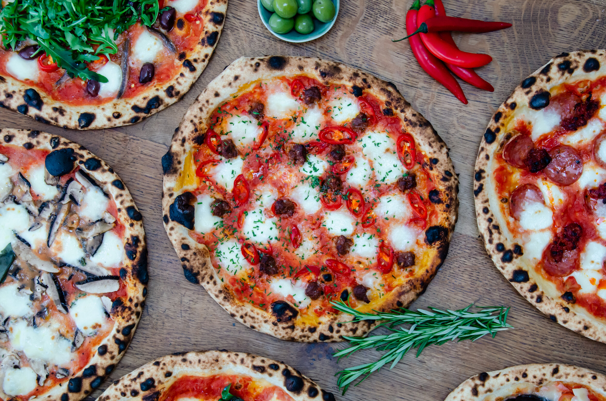 Various wood-fired pizzas topped with ingredients like cheese, mushrooms, tomatoes, arugula, and red chili peppers, surrounded by rosemary, green olives, and whole red chili peppers on a wooden surface.