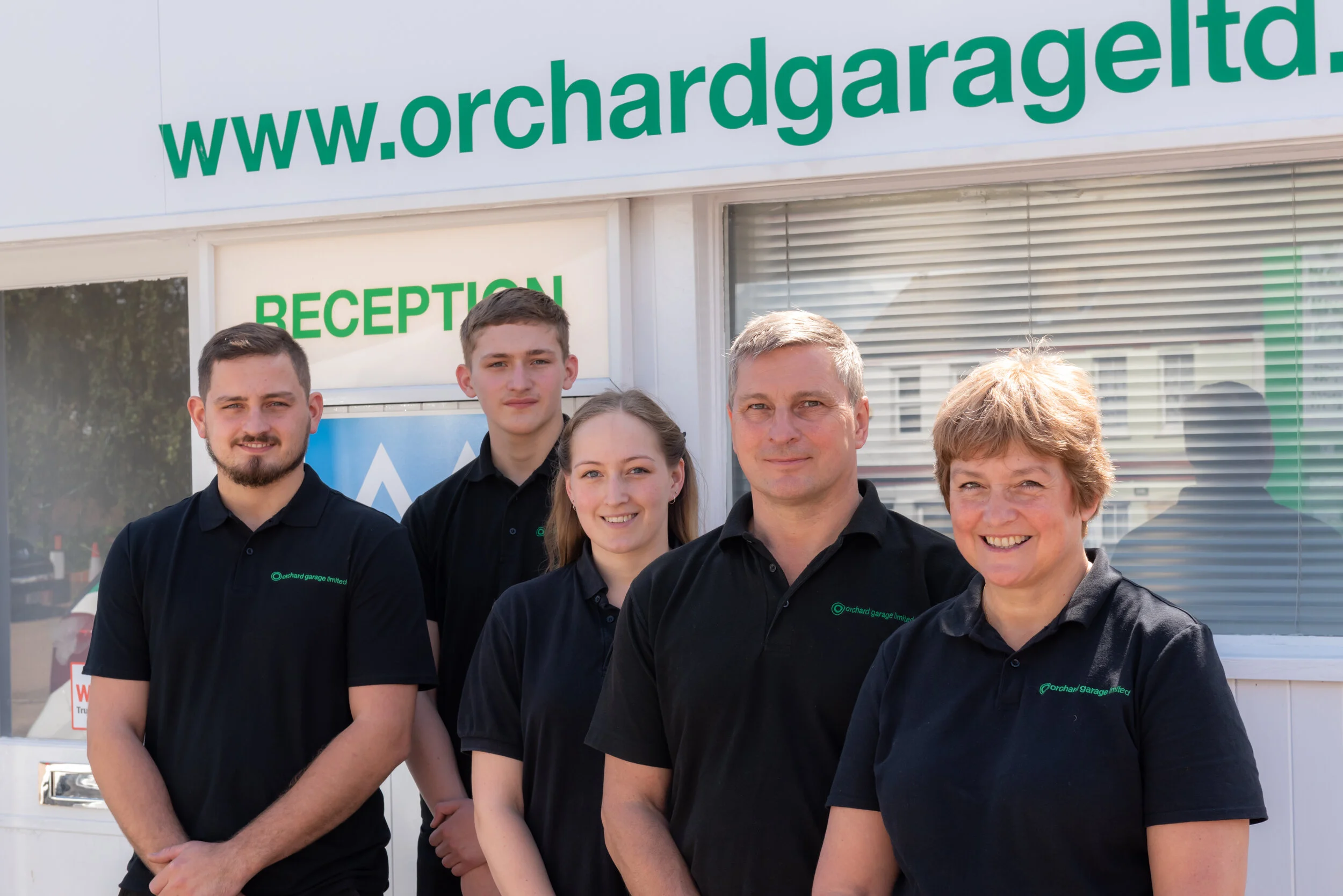 Group of five people in black polo shirts standing outside a building with a sign "www.orchardgarageltd.co.uk" and "Reception."