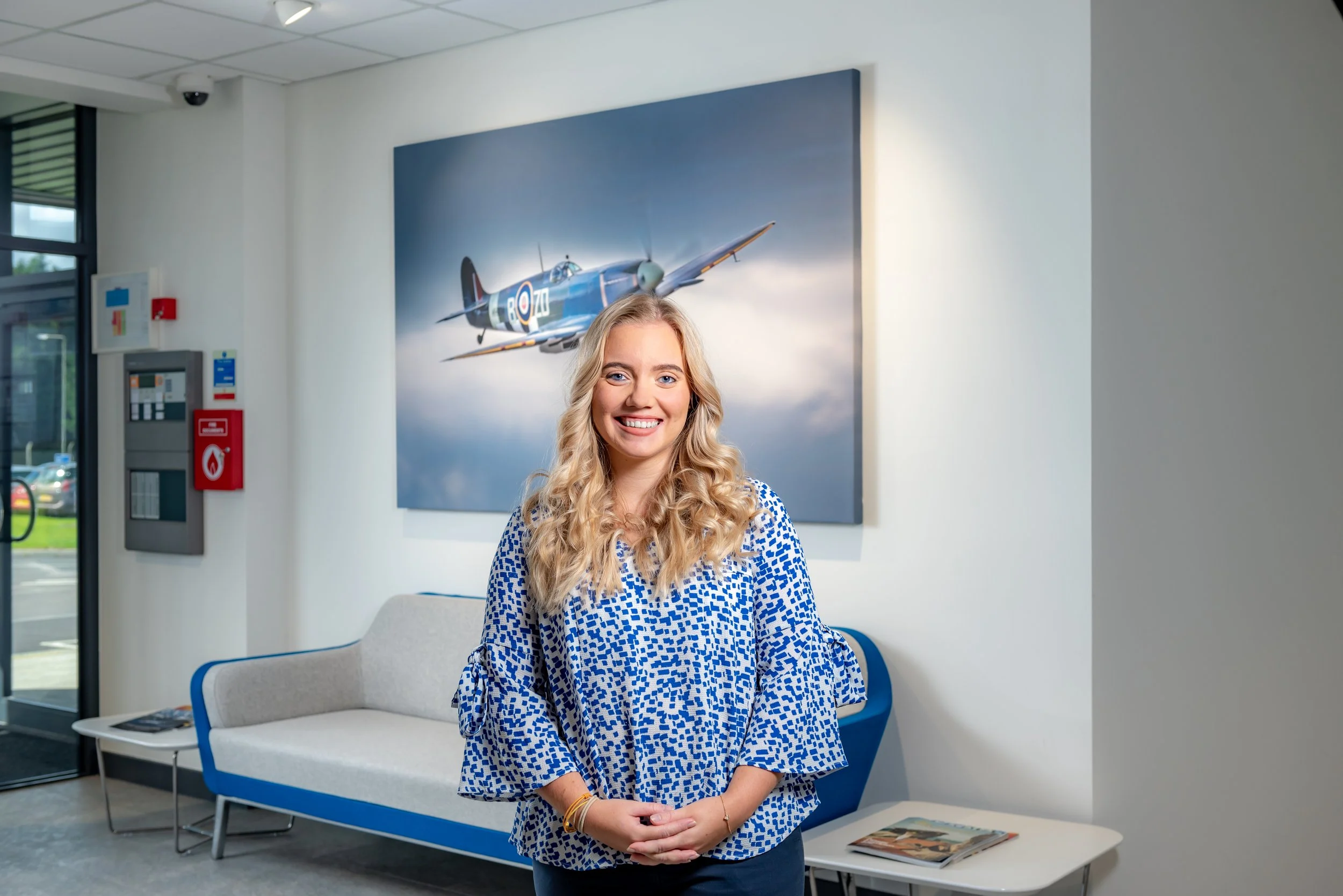 Smiling woman standing in an office lobby in front of a large painting of a historical aircraft on a wall, next to a sofa and a small table with magazines.