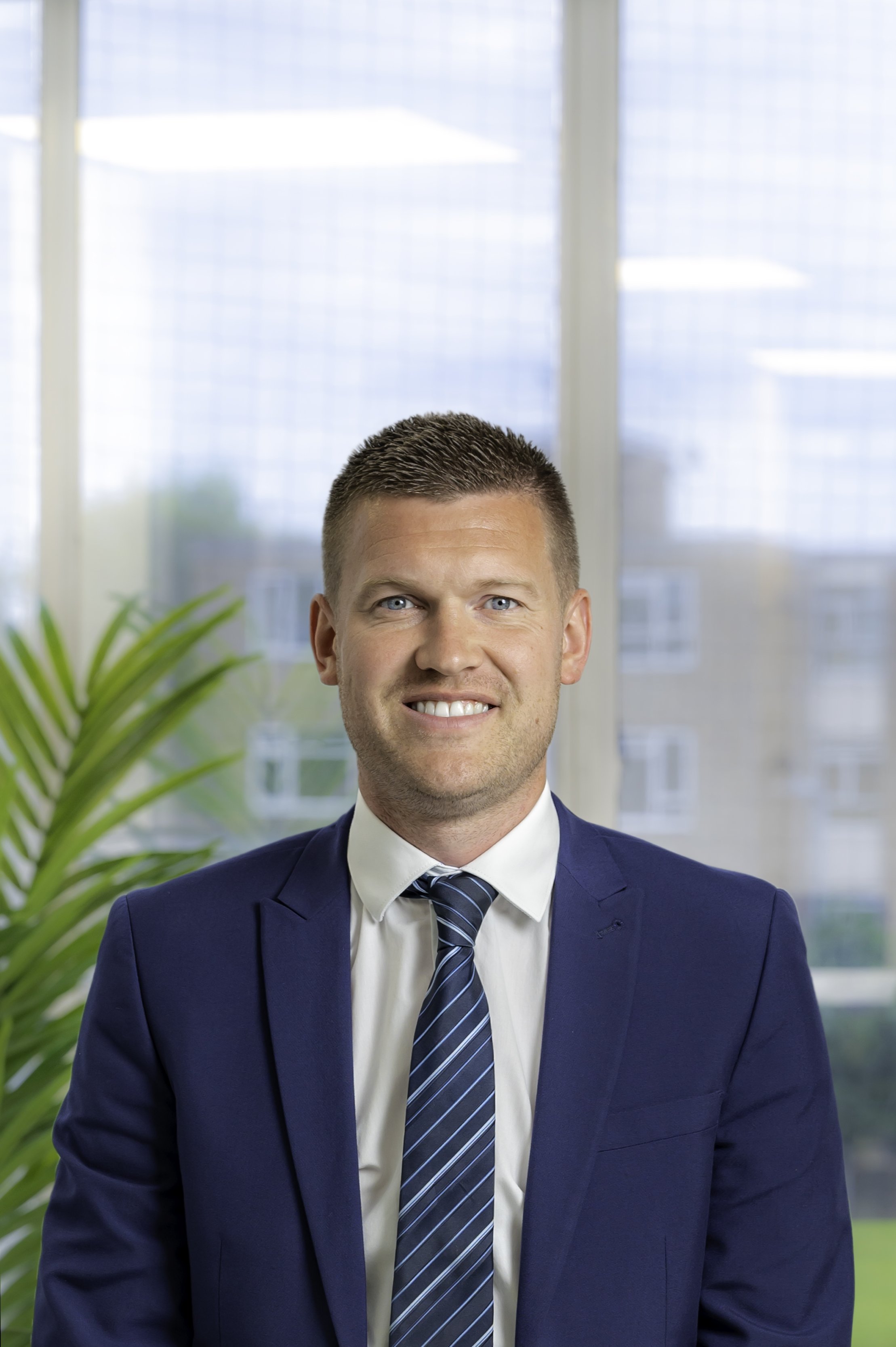 Man in blue suit and striped tie smiling in an office with a plant and window.