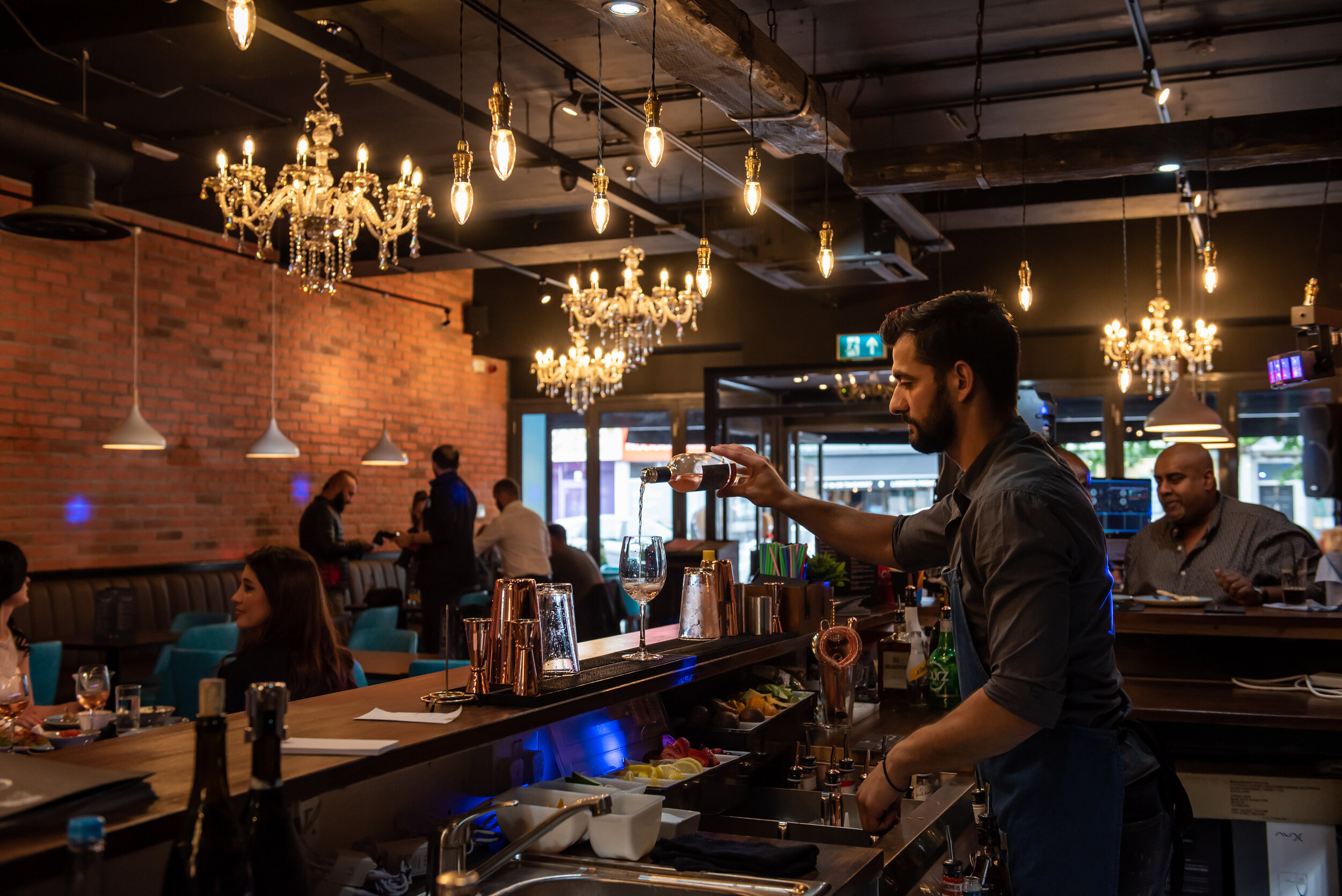 Chic bar interior with bartender pouring wine, stylish chandeliers, exposed brick walls, and patrons seated at tables.