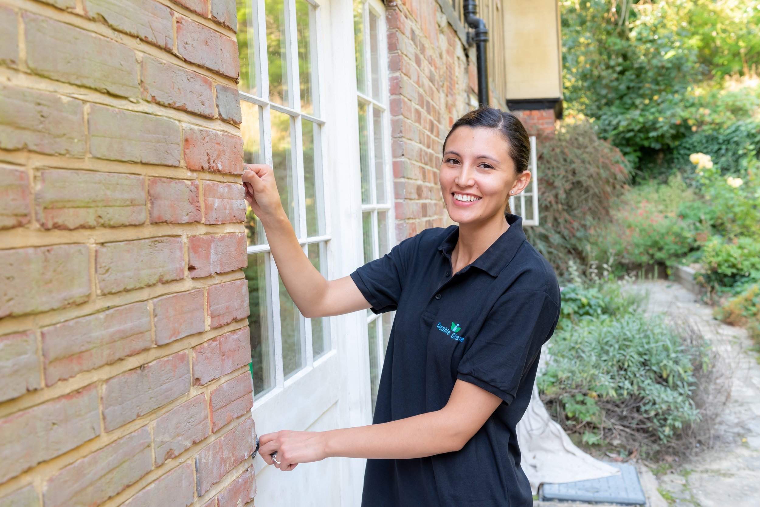 Woman smiling, unlocking a door with a key, wearing a black polo shirt, standing outside a brick building with a garden.