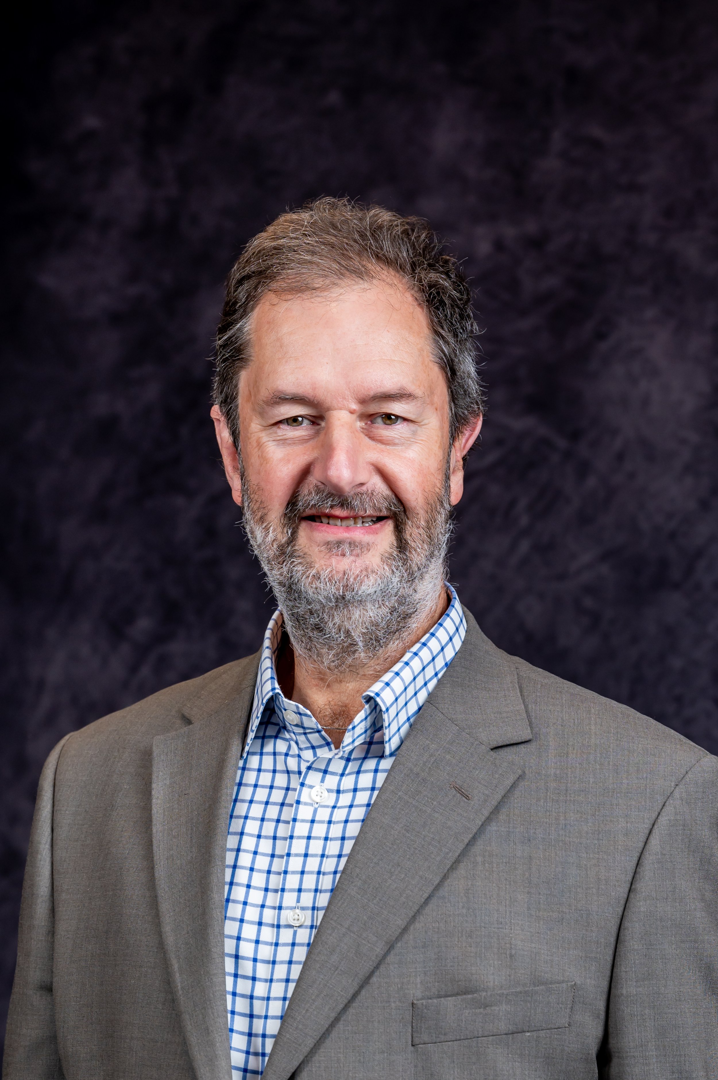 Smiling man with a beard in a suit and checkered shirt, dark background.