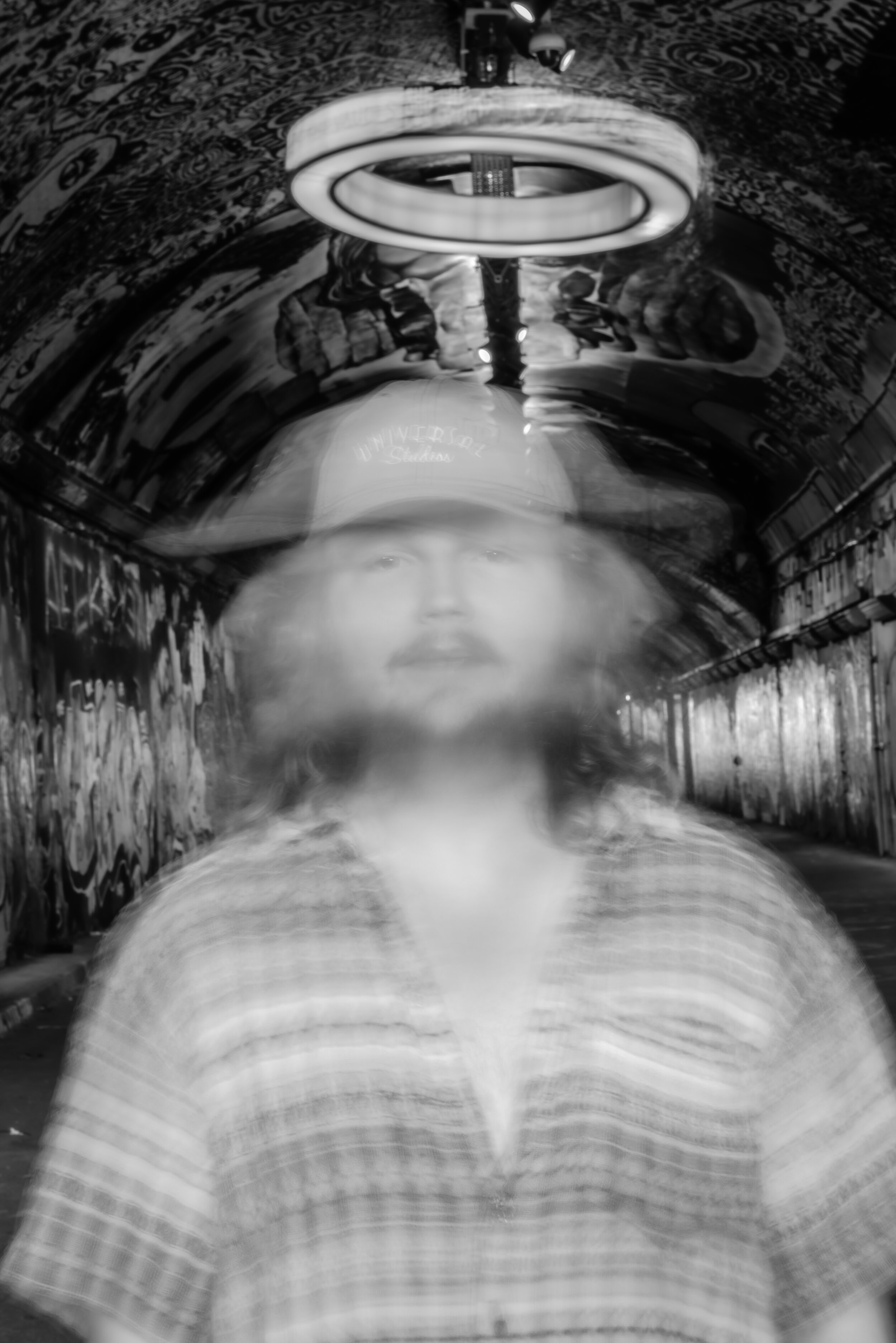Blurred black and white image of a person wearing a plaid shirt and cap, standing in a graffiti-covered tunnel.
