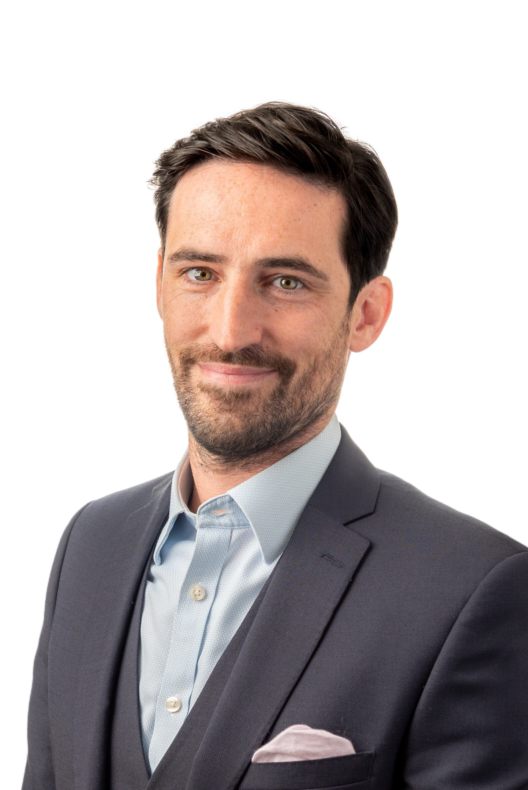 Man with dark hair wearing a suit and light blue dress shirt, smiling, against a white background.