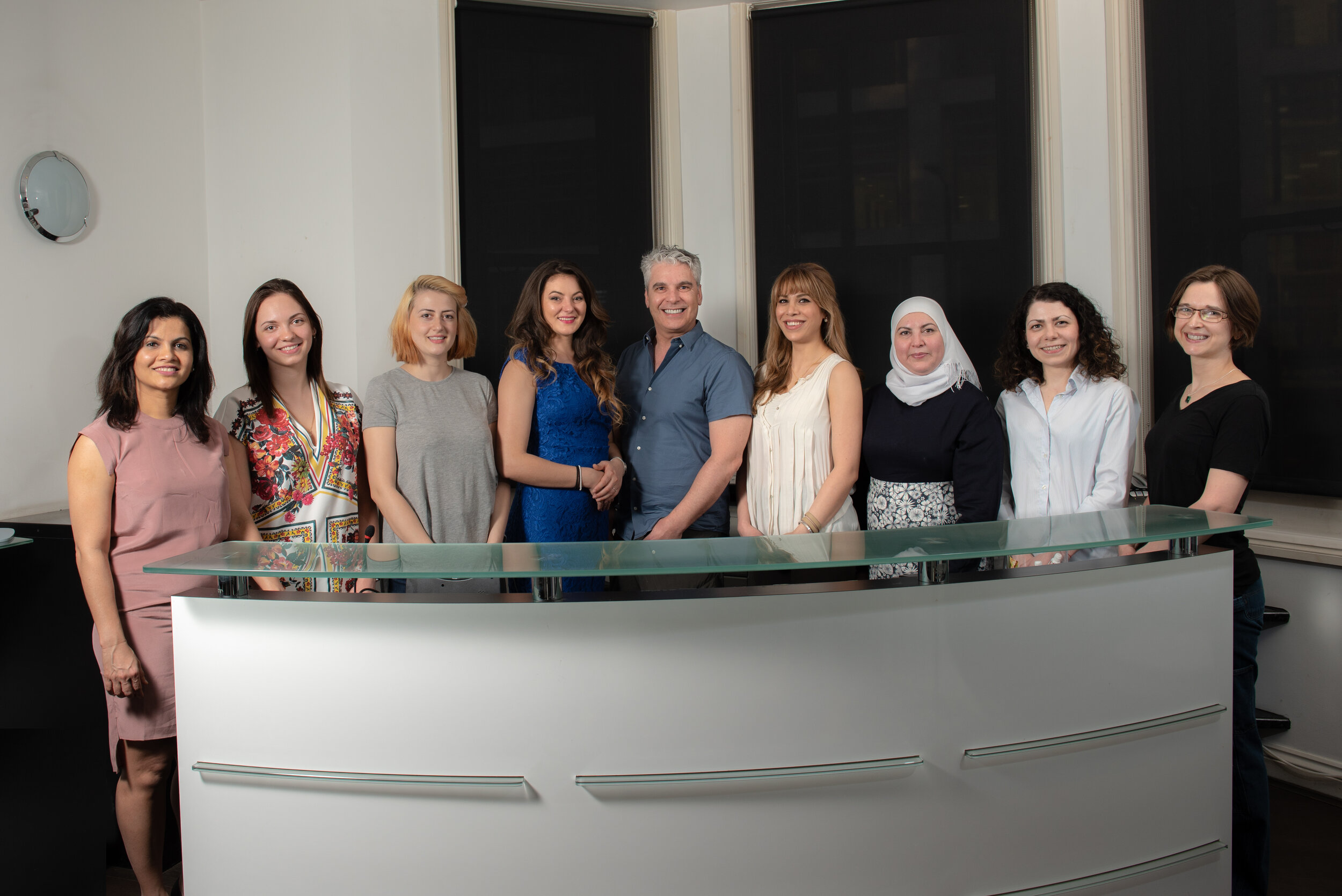 Group of ten people standing behind a modern reception desk in an office setting; background has black window blinds.
