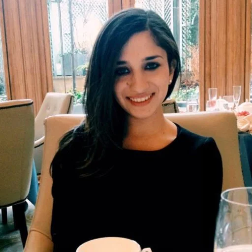 Woman with long dark hair wearing a black top, smiling while seated at a dining table with a window and curtains in the background.